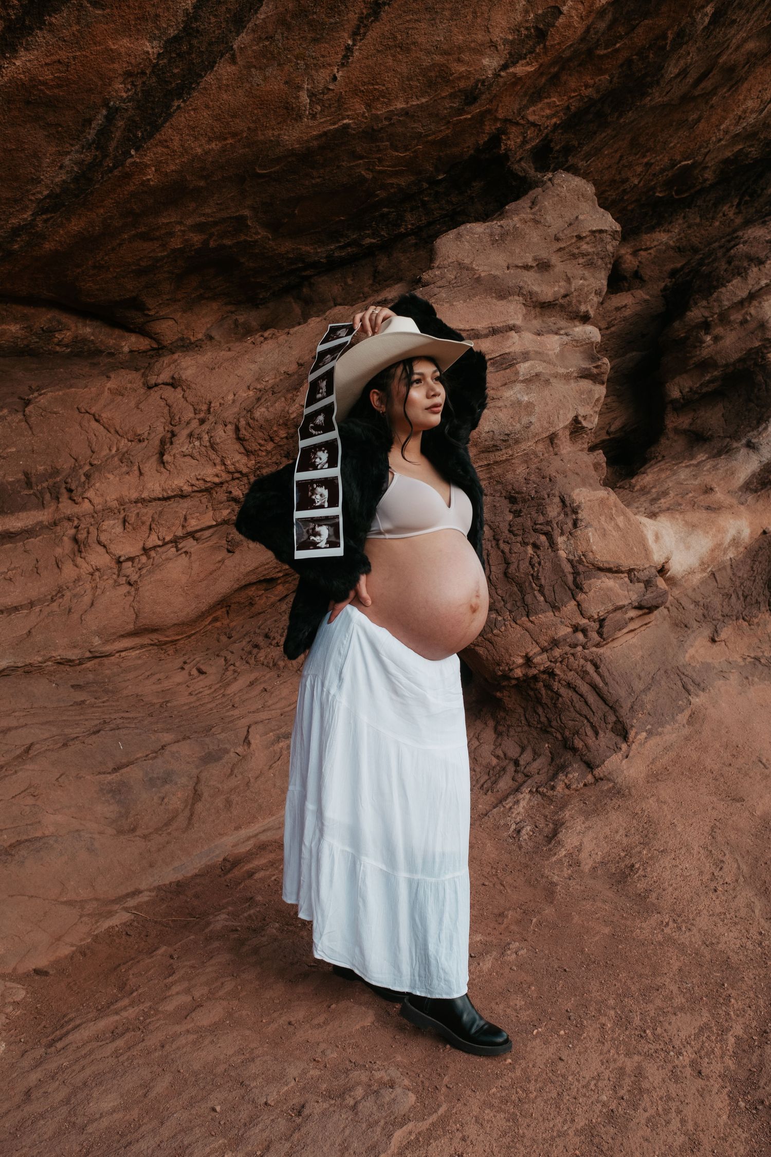 Pregnant woman in cowboy hat holds ultrasound strip against red sandstone rock formation.