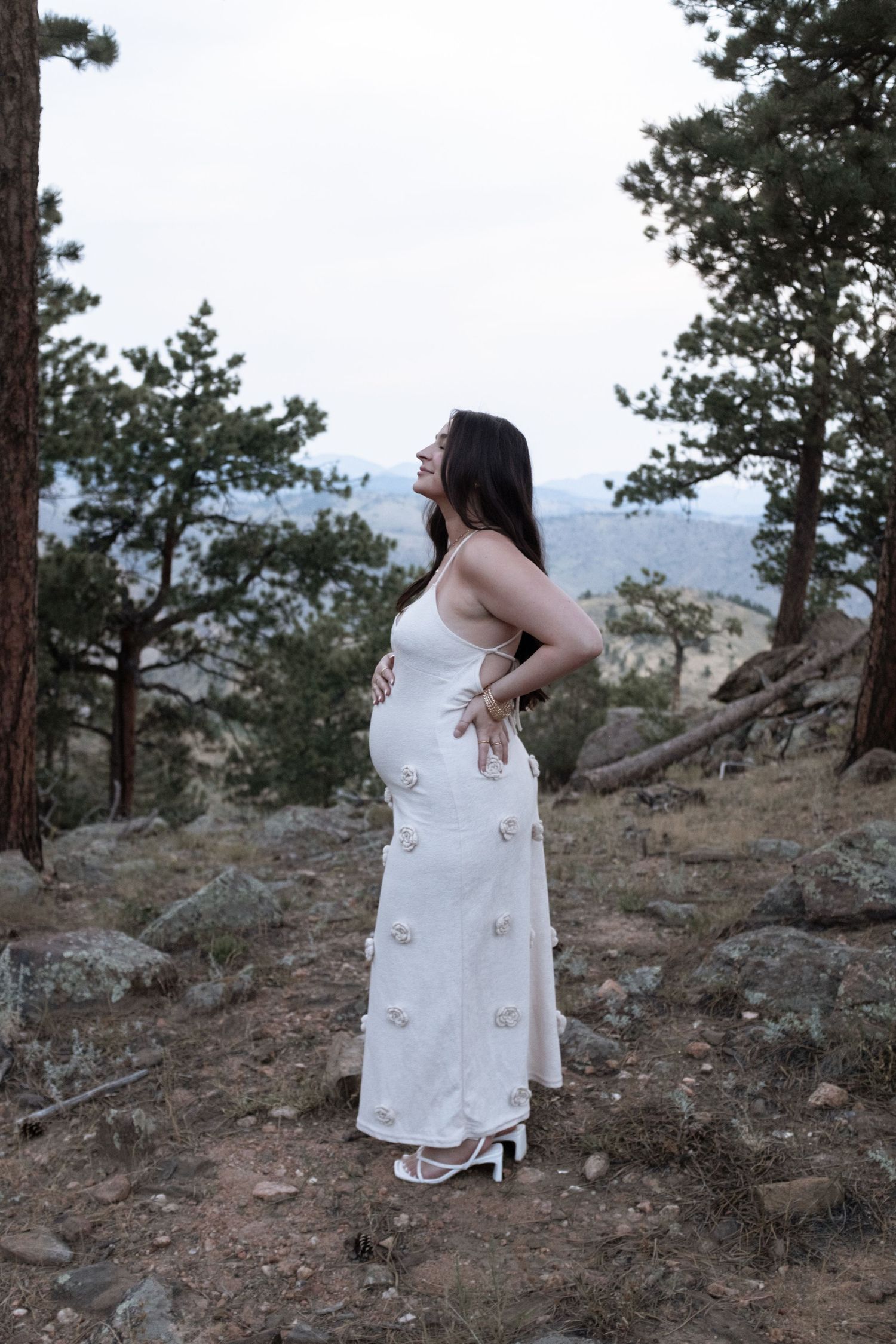 Pregnant woman in white floral maxi dress standing outdoors among pine trees on rocky hillside, cradling baby bump.
