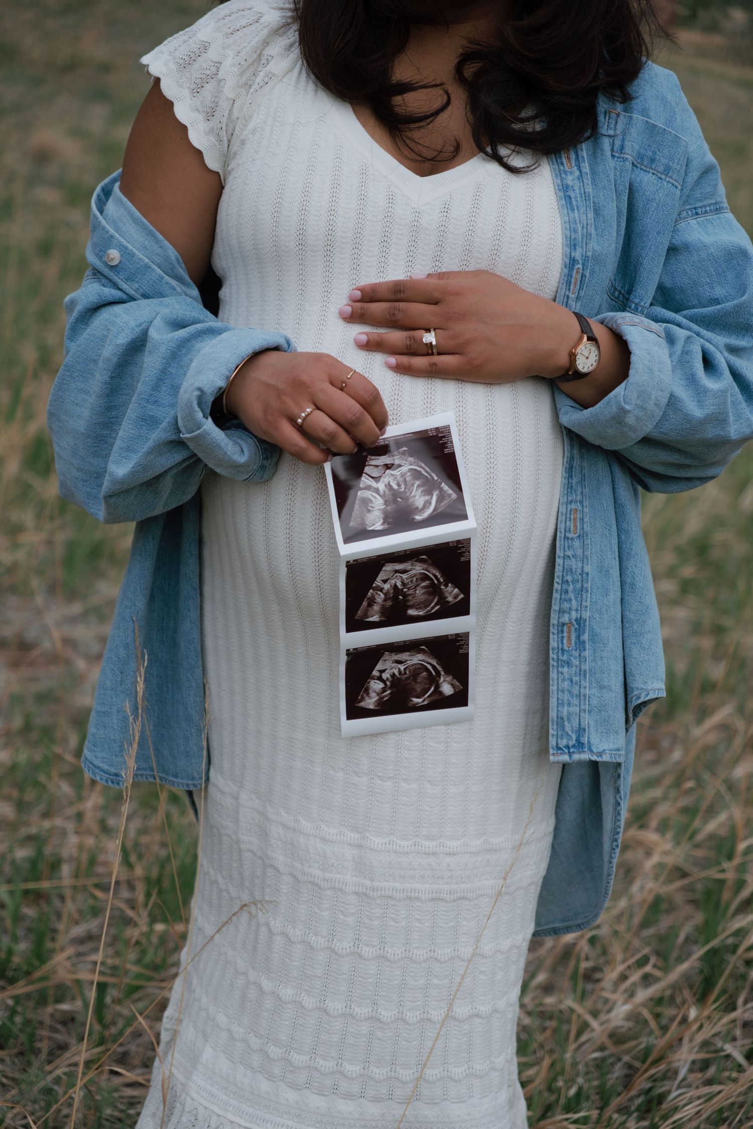 Pregnant woman in white dress and denim jacket holding ultrasound photos against her baby bump outdoors.