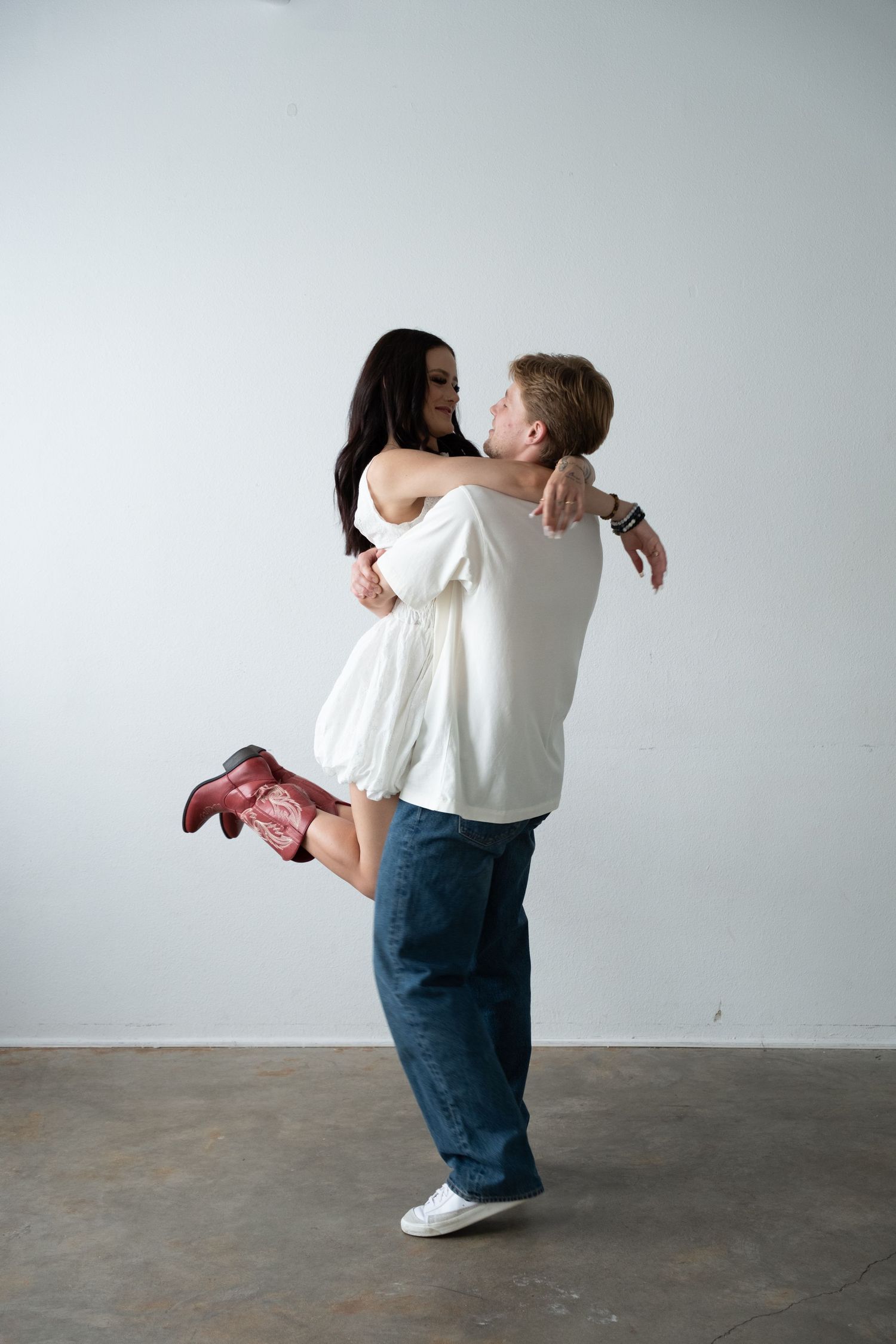Man in white shirt lifting woman in white dress and red boots, sharing a romantic embrace against white wall.