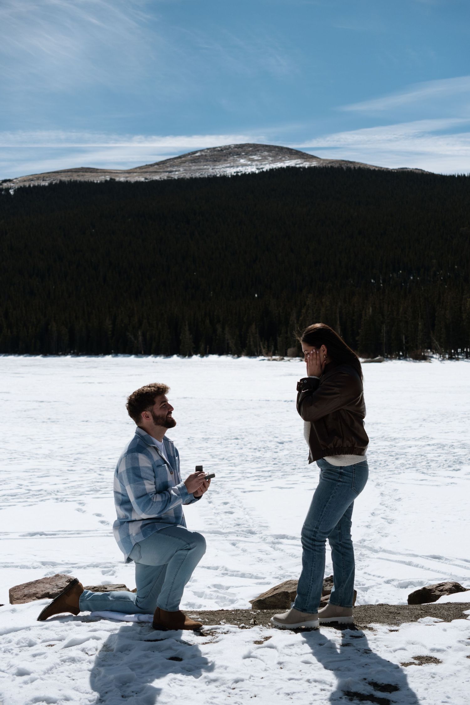 Man proposes on one knee to woman in snowy mountain landscape with frozen lake and pine forest backdrop.