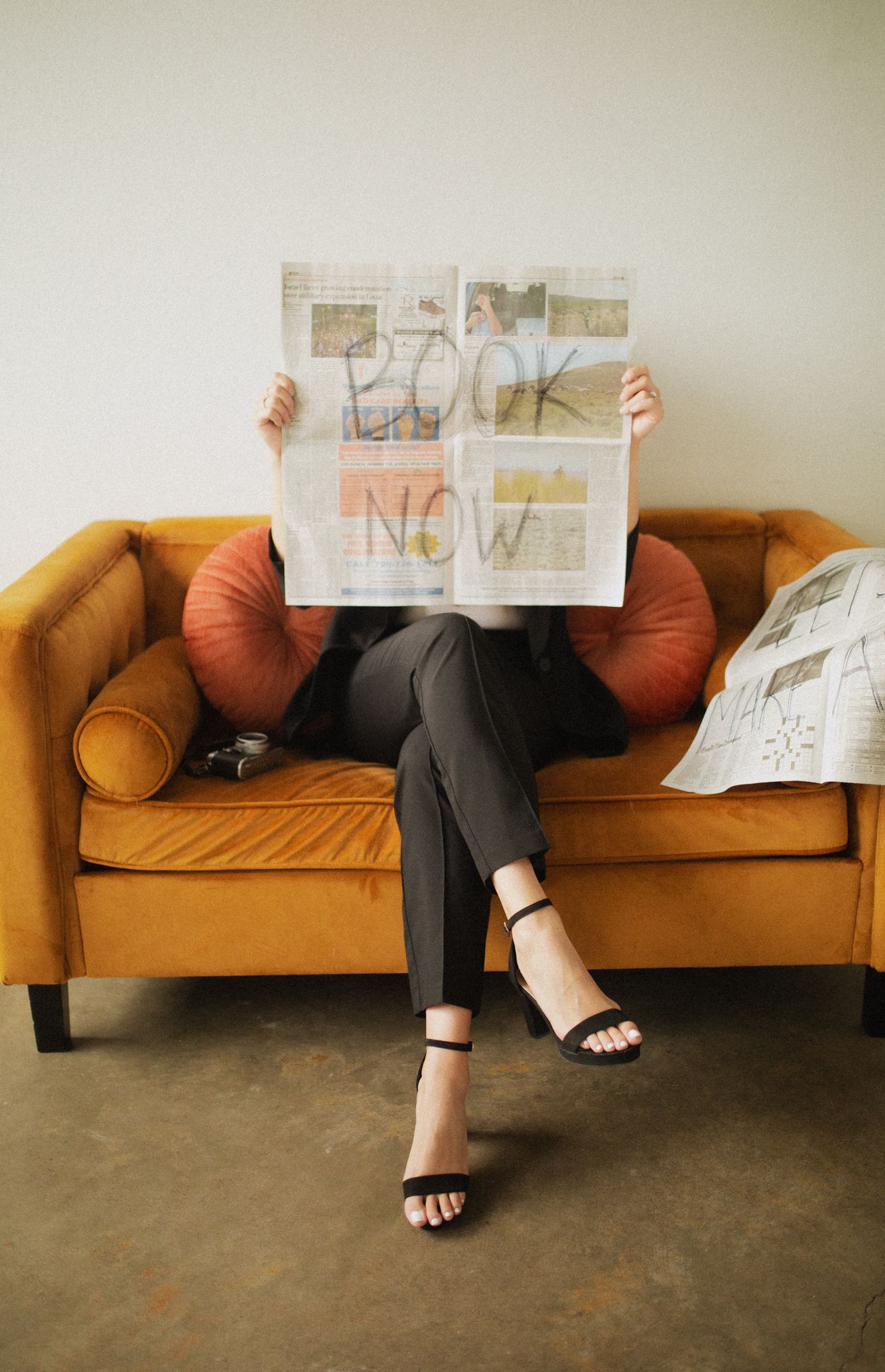 Person sitting on vintage orange velvet couch reading newspaper with stylish black sandals.