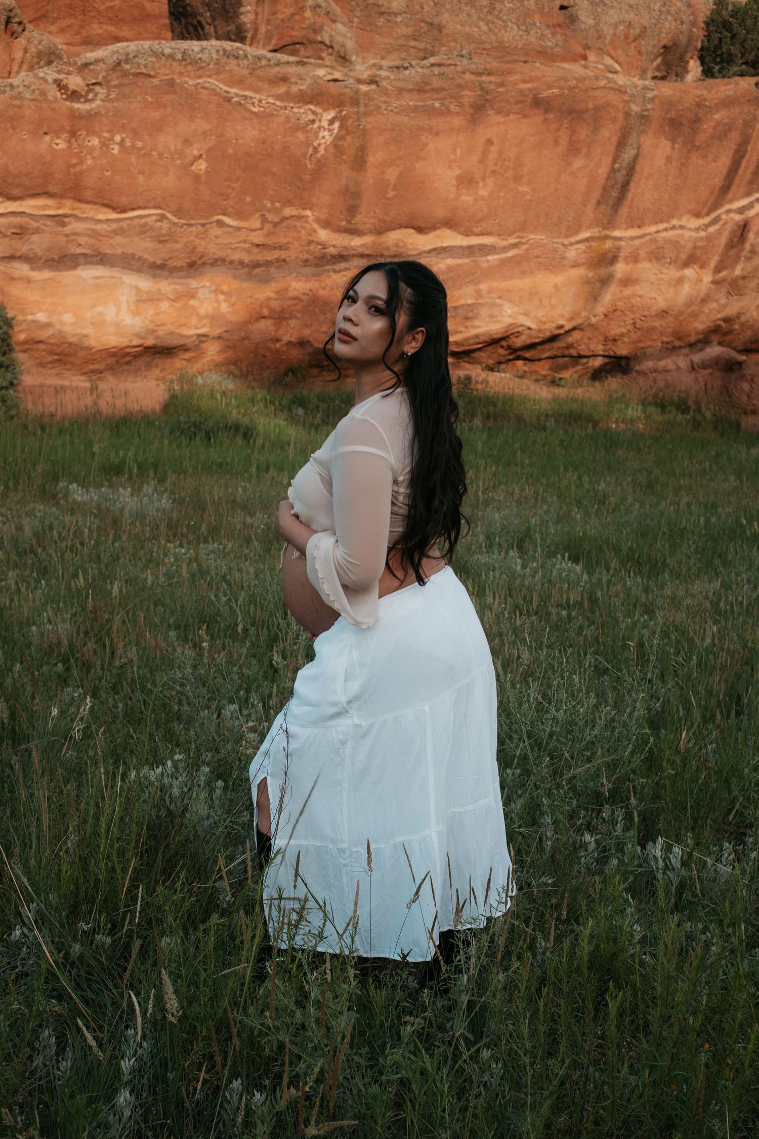 Side profile maternity shot with warm red rock formations and tall grass.