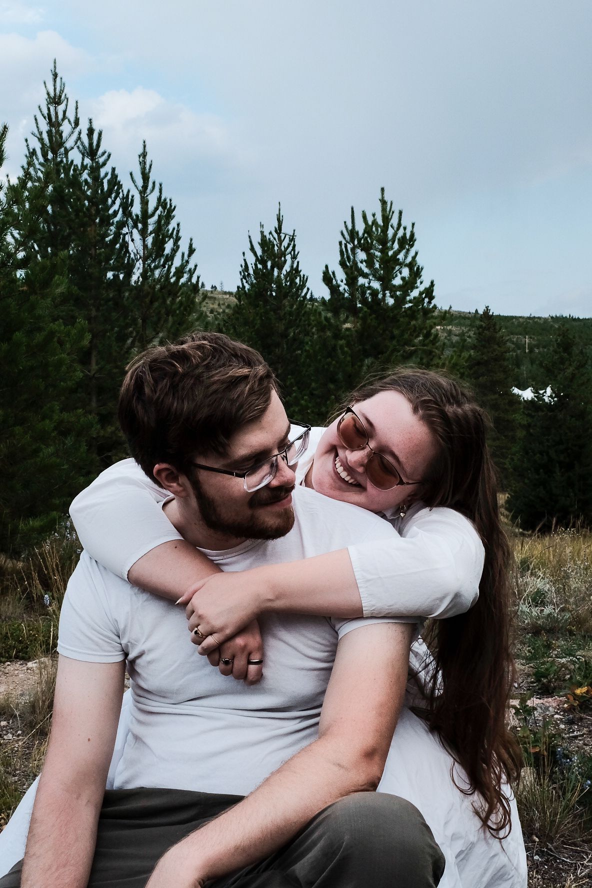 Couple in white shirts share intimate embrace against backdrop of pine trees.