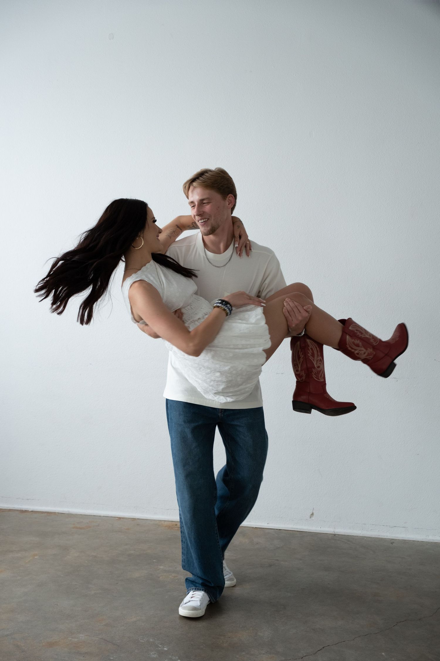 A man in jeans and white tee carries a smiling woman in red cowboy boots, both gazing at each other happily.