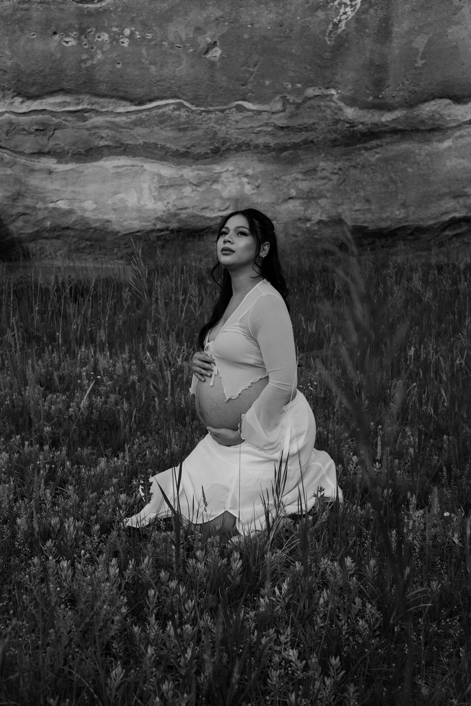 Black and white maternity photo of pregnant woman kneeling in field with rocky cliff backdrop.