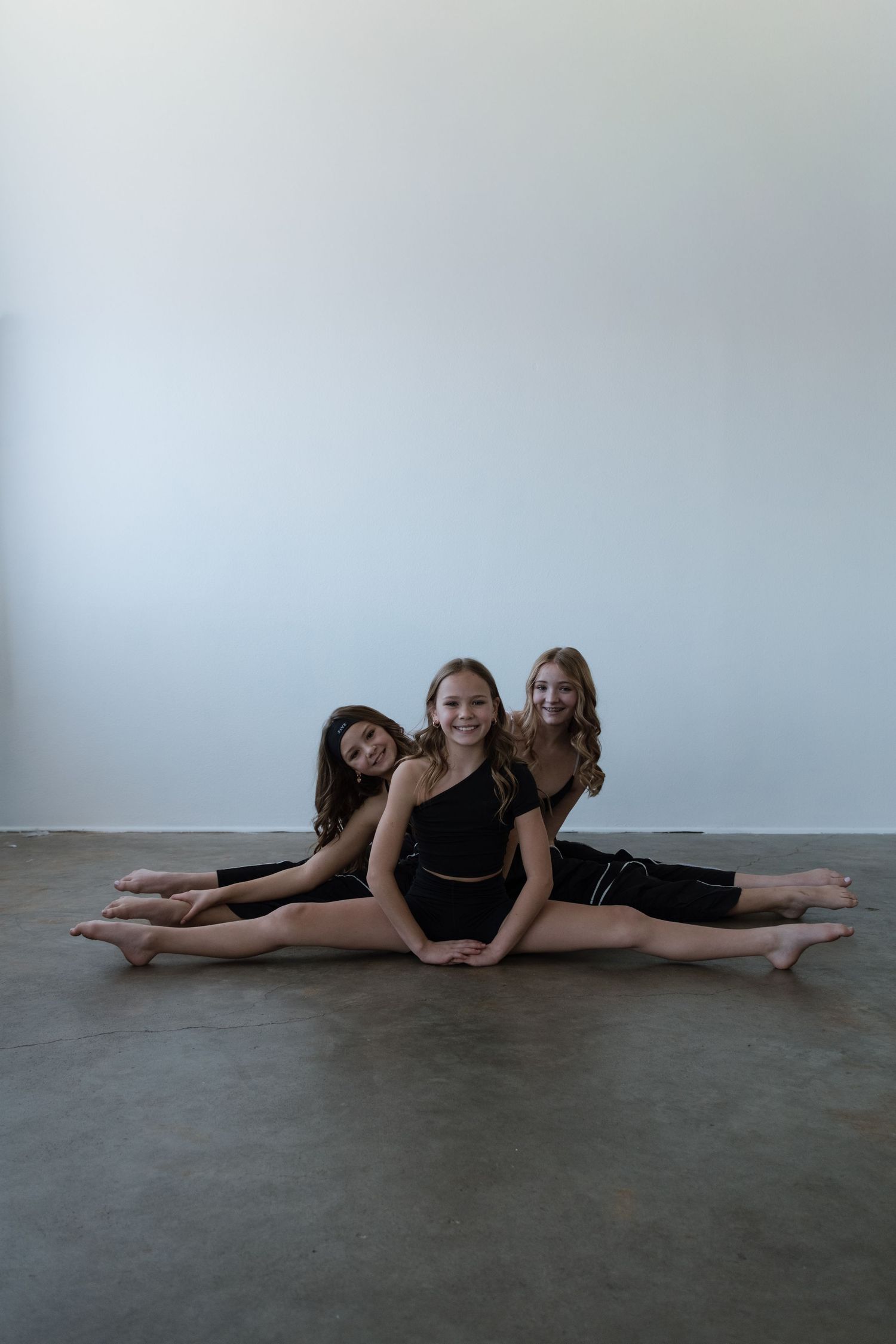 Three young dancers in black outfits perform splits on studio floor, smiling at camera.