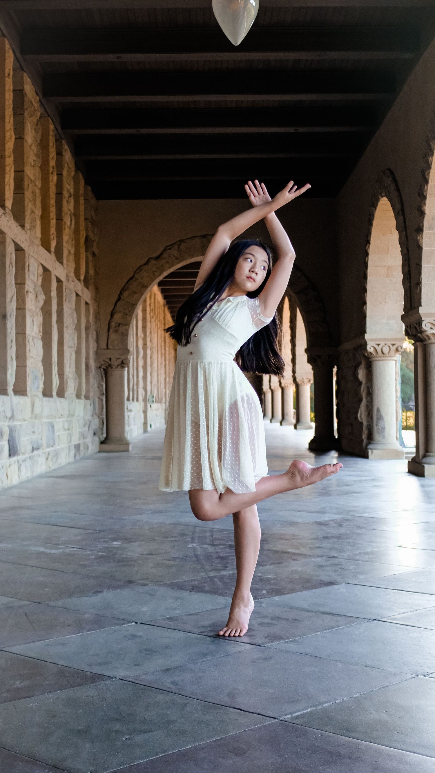 Dancer in white dress performs expressive movement in arched stone hallway.