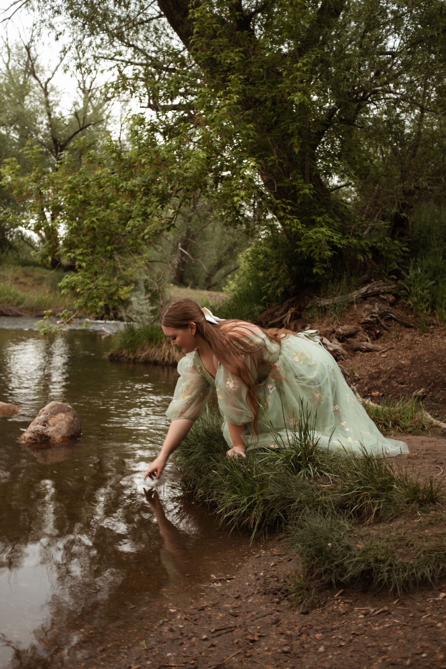Figure in flowing dress leans down to touch stream water in serene forest setting.