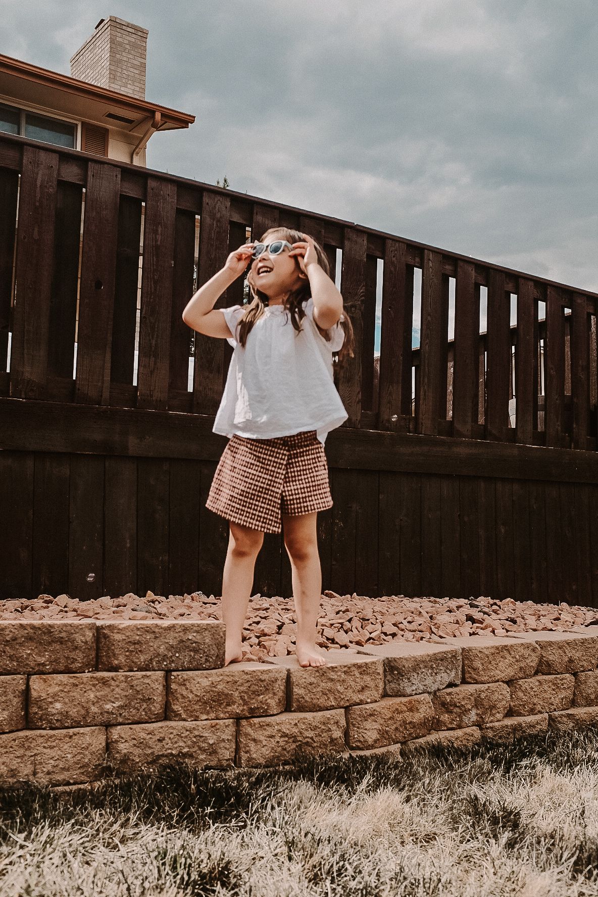 A child in summer clothes stands on brick retaining wall against wooden fence backdrop.