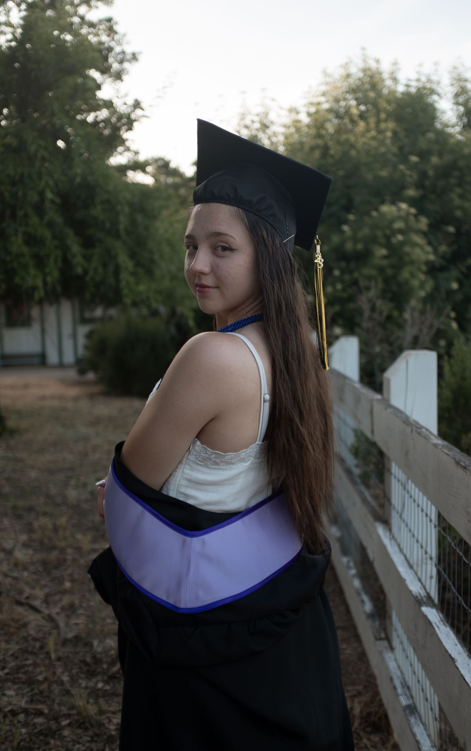Graduate in traditional cap and gown poses by white fence at sunset.