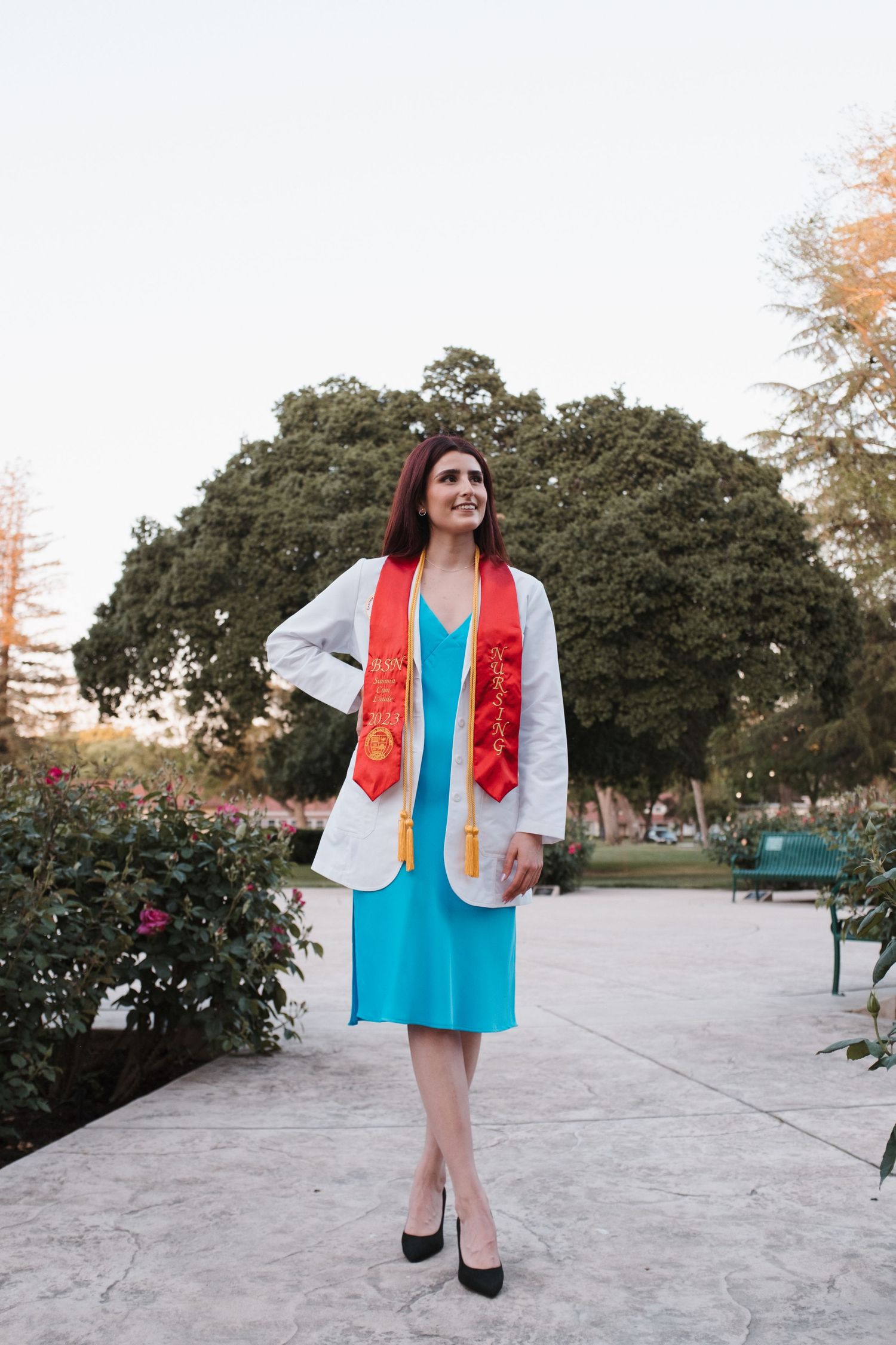 Graduate wearing turquoise dress and red stole poses outdoors in park setting.