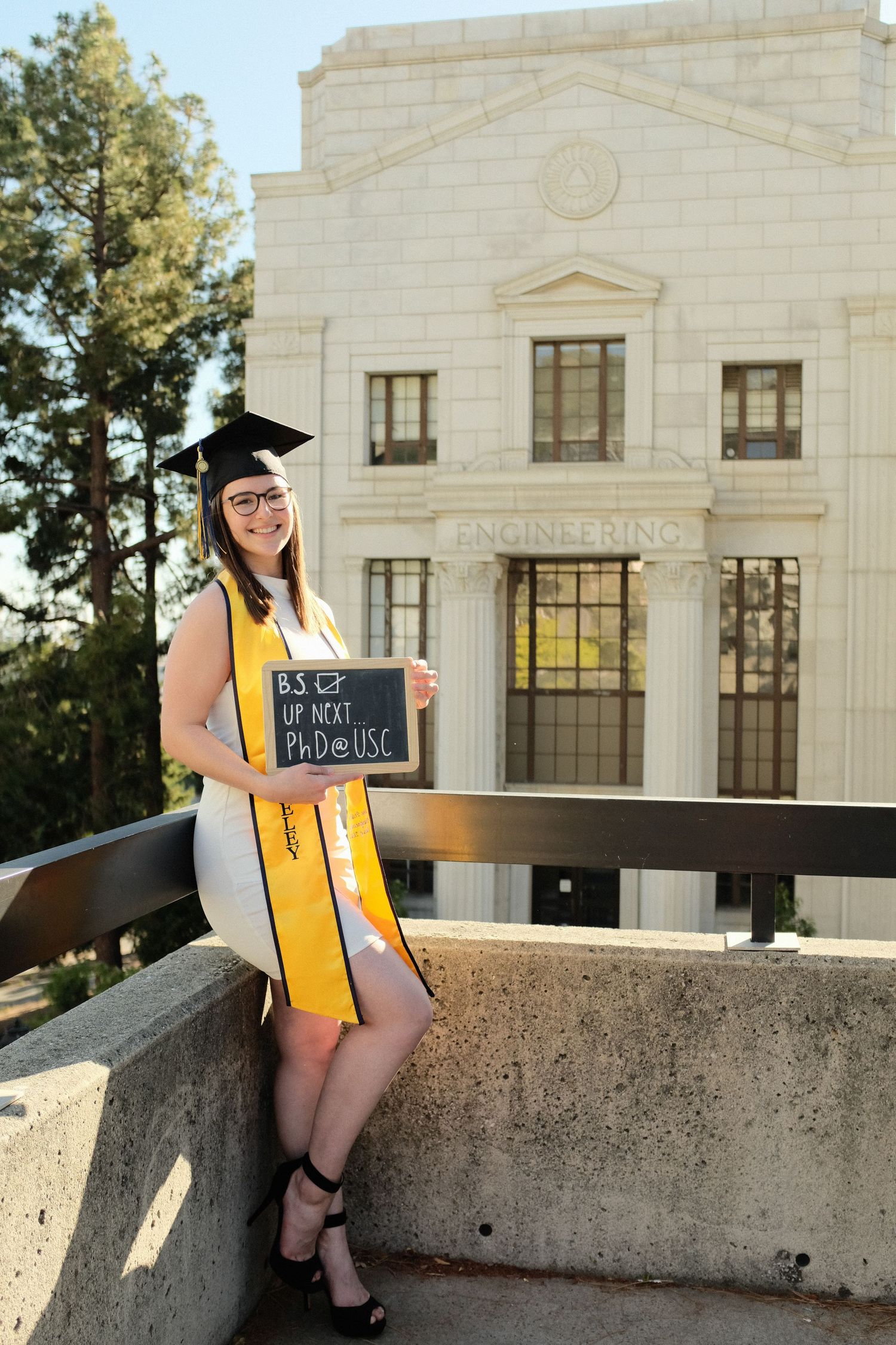 A graduate in yellow and white stole stands outside a stone building holding a sign on a sunny day.