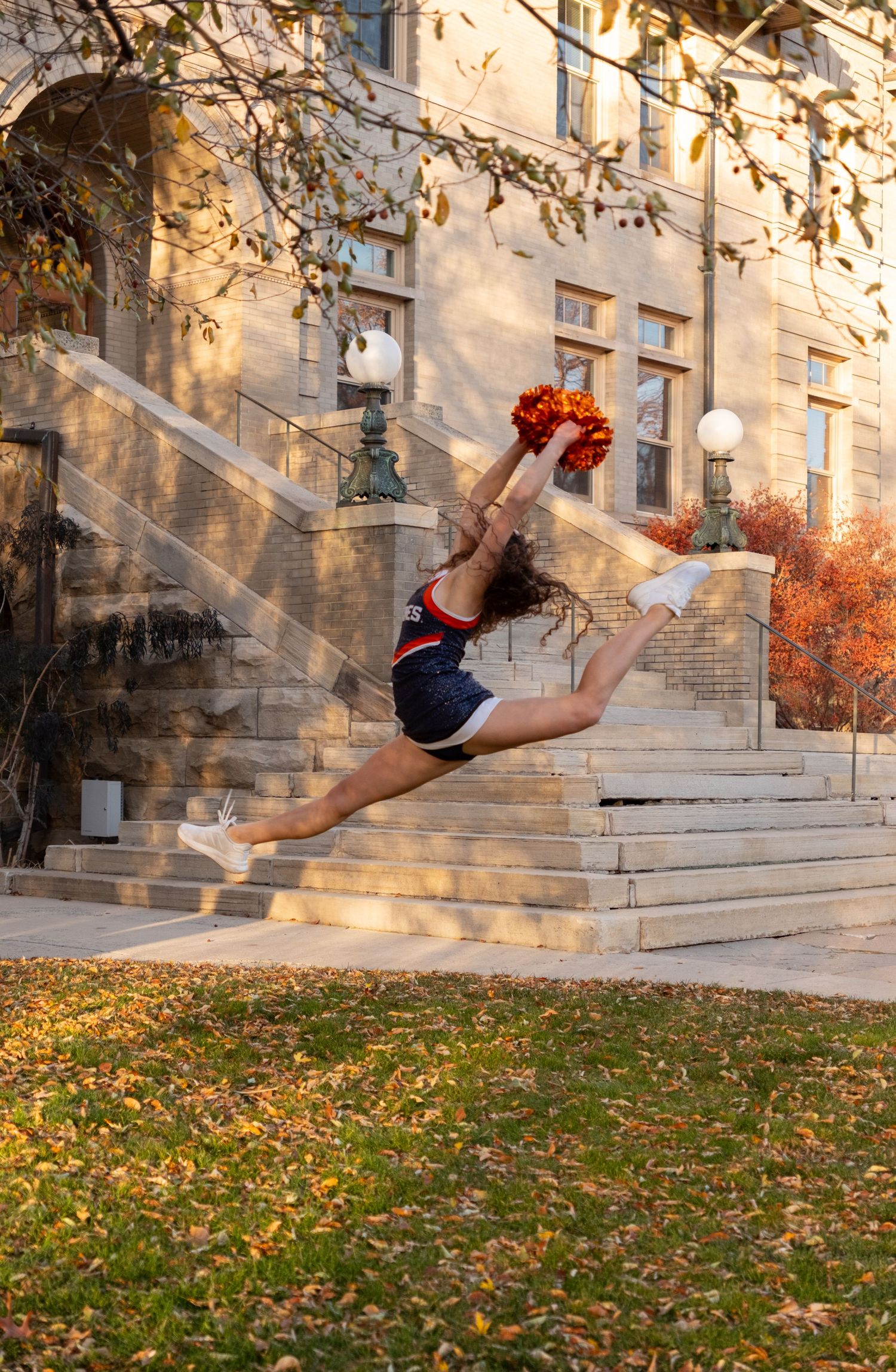 Cheerleader in navy uniform leaps mid-air with orange pom-poms outside a stone building surrounded by autumn foliage.