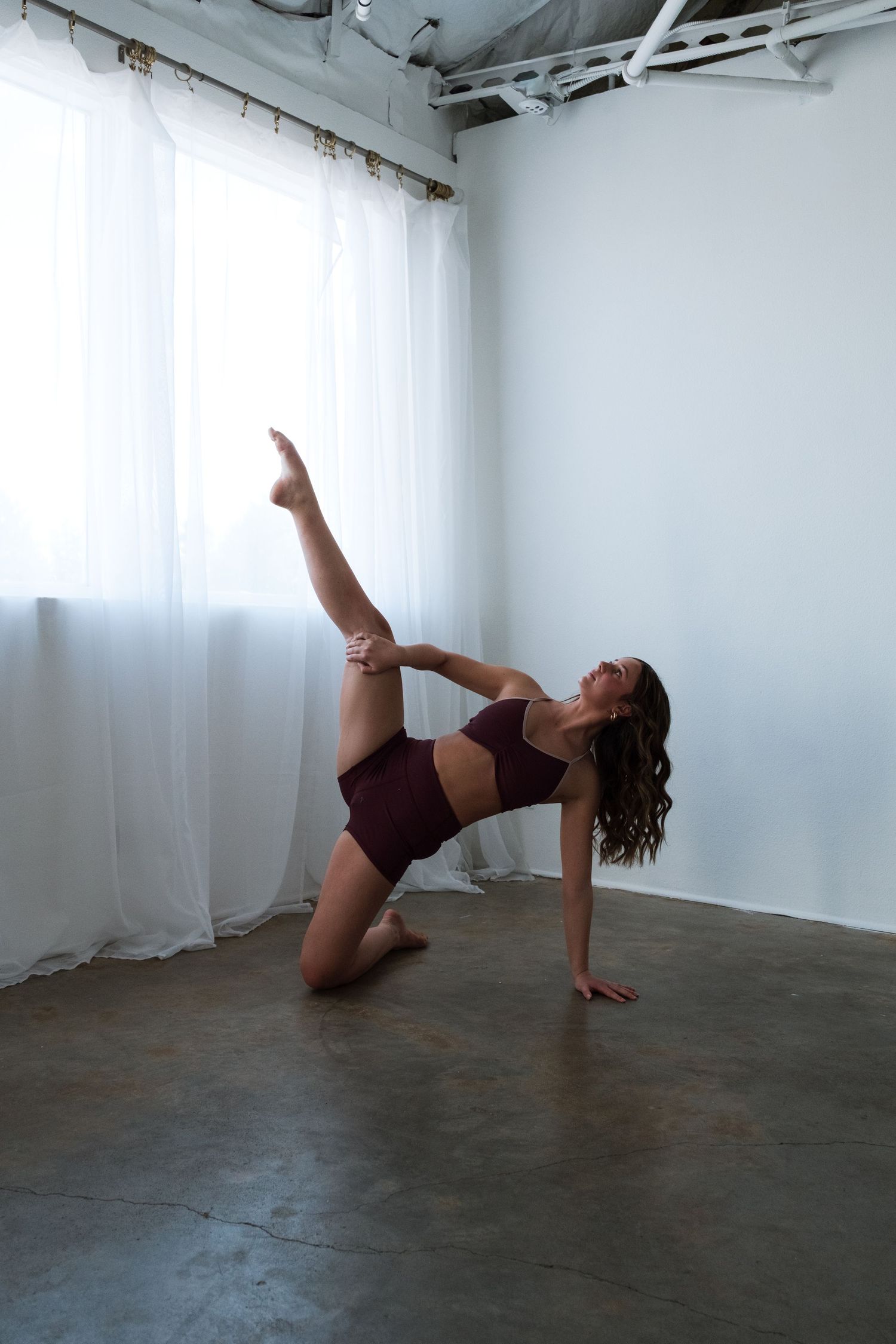 Flexible dancer in maroon activewear performing a side balance pose with leg raised in a bright white studio.
