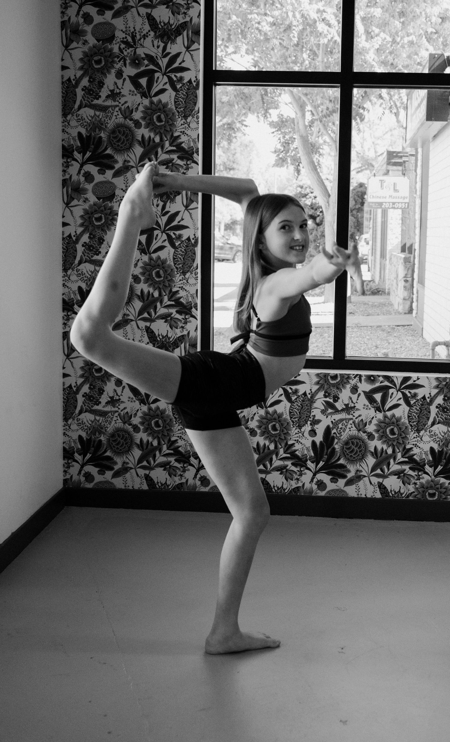 Young dancer performs a graceful standing split pose in a dance studio with floral wallpaper, black and white photo.