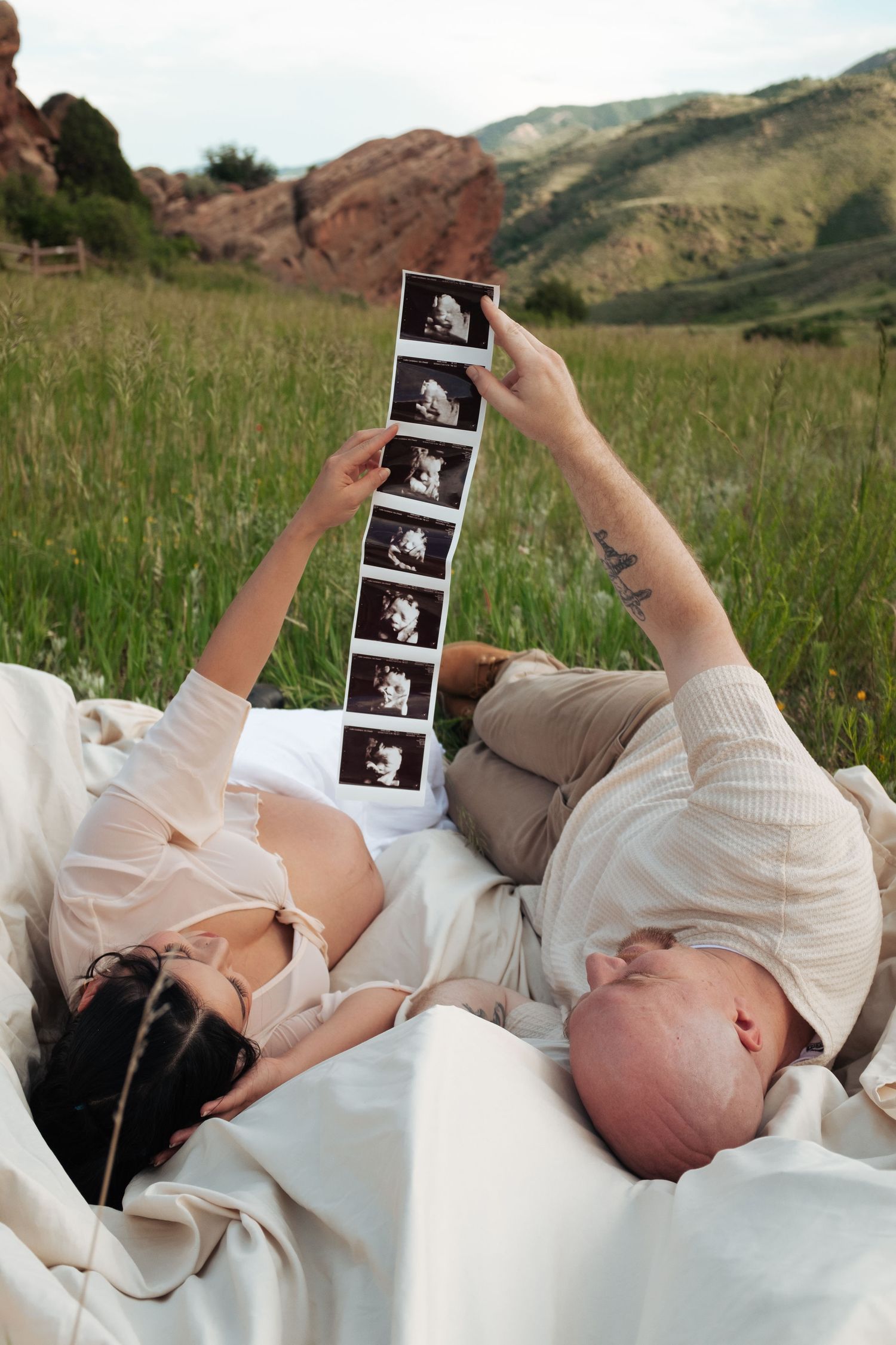Expectant couple lies on blanket in field viewing ultrasound images with red rocks behind them.