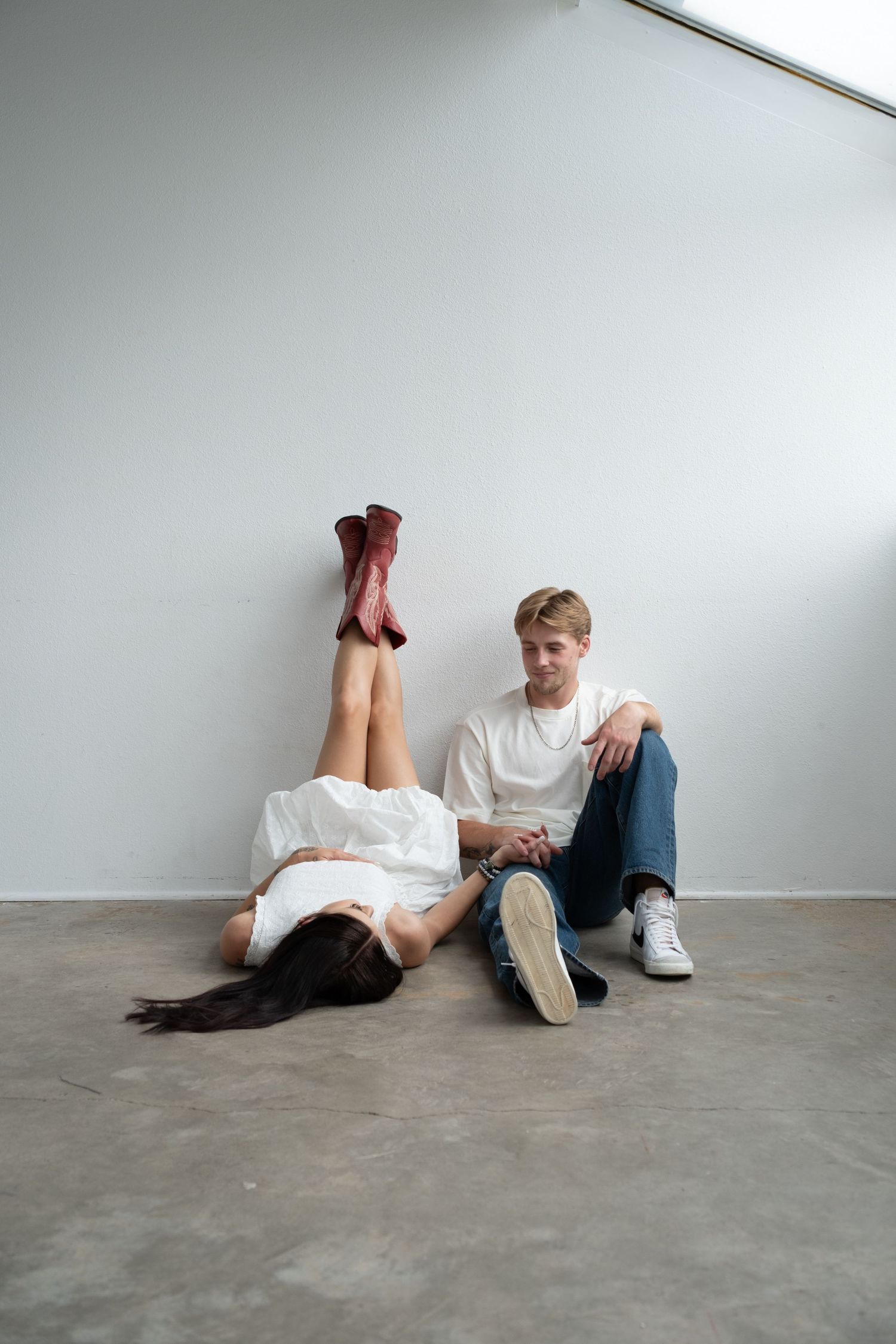 Young couple sitting on floor against white wall, woman lying down with legs up, man in white tee and jeans looking at her.