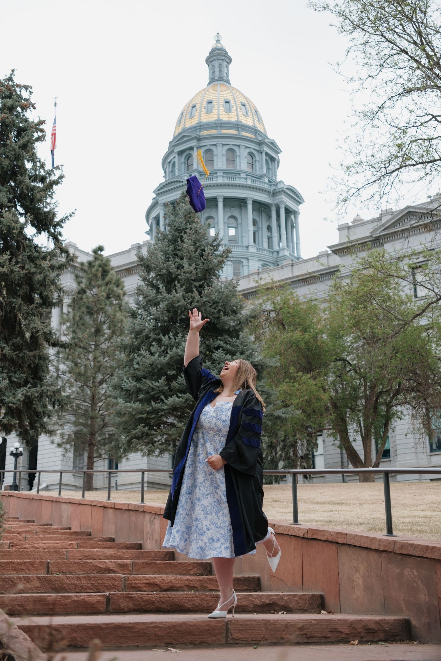 Graduate in black gown tosses cap in front of Colorado State Capitol building with golden dome.