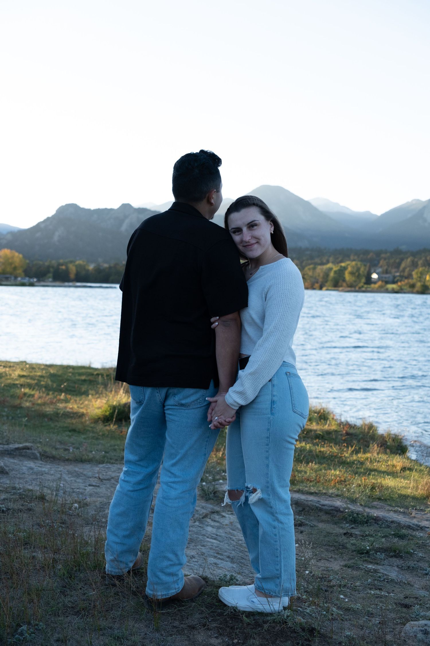 Couple holding hands by a mountain lake at sunset, woman in white sweater smiling at camera, man in black shirt facing water.