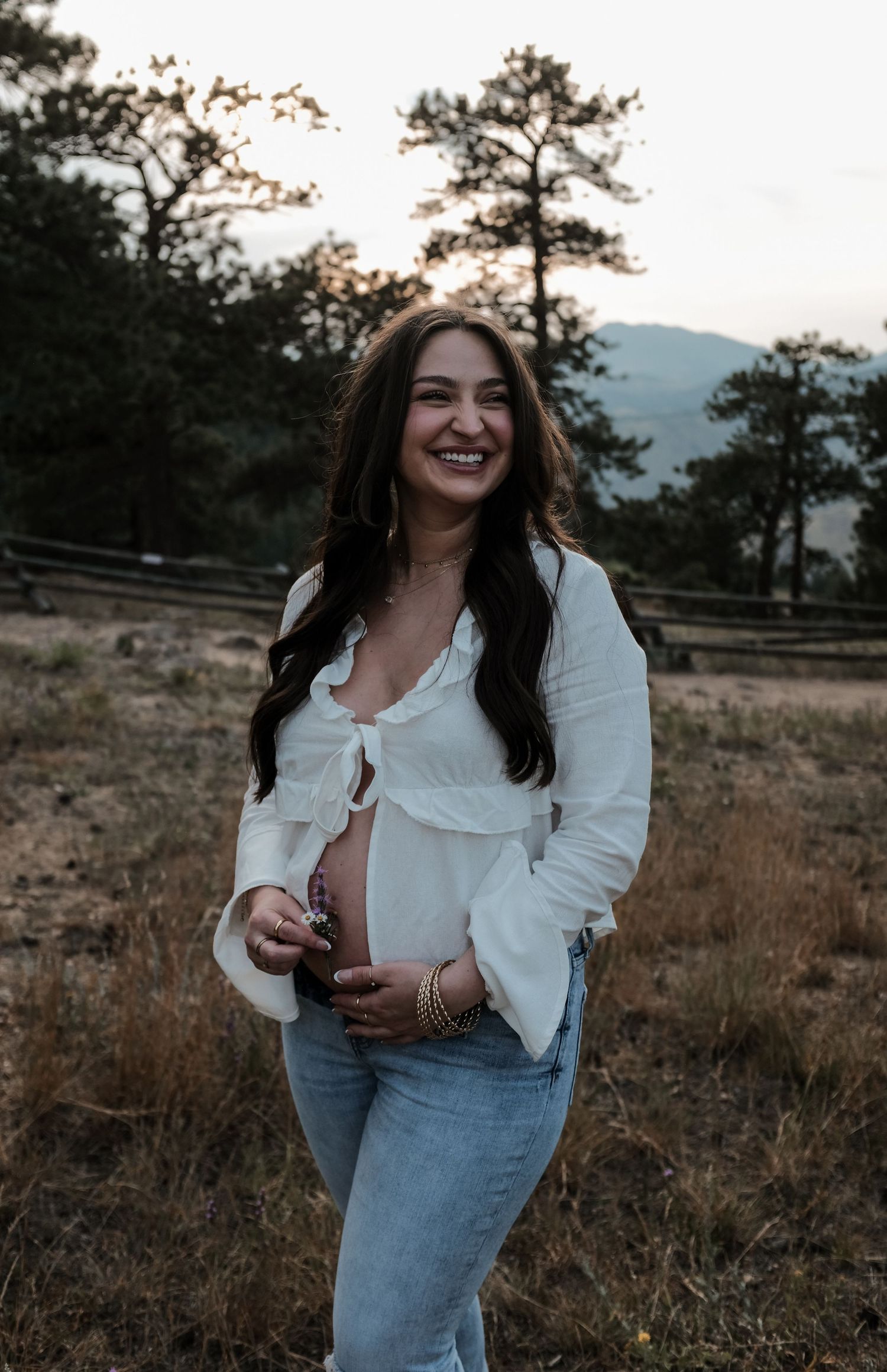 Smiling pregnant woman in white top and jeans poses outdoors at golden hour with mountains in background.