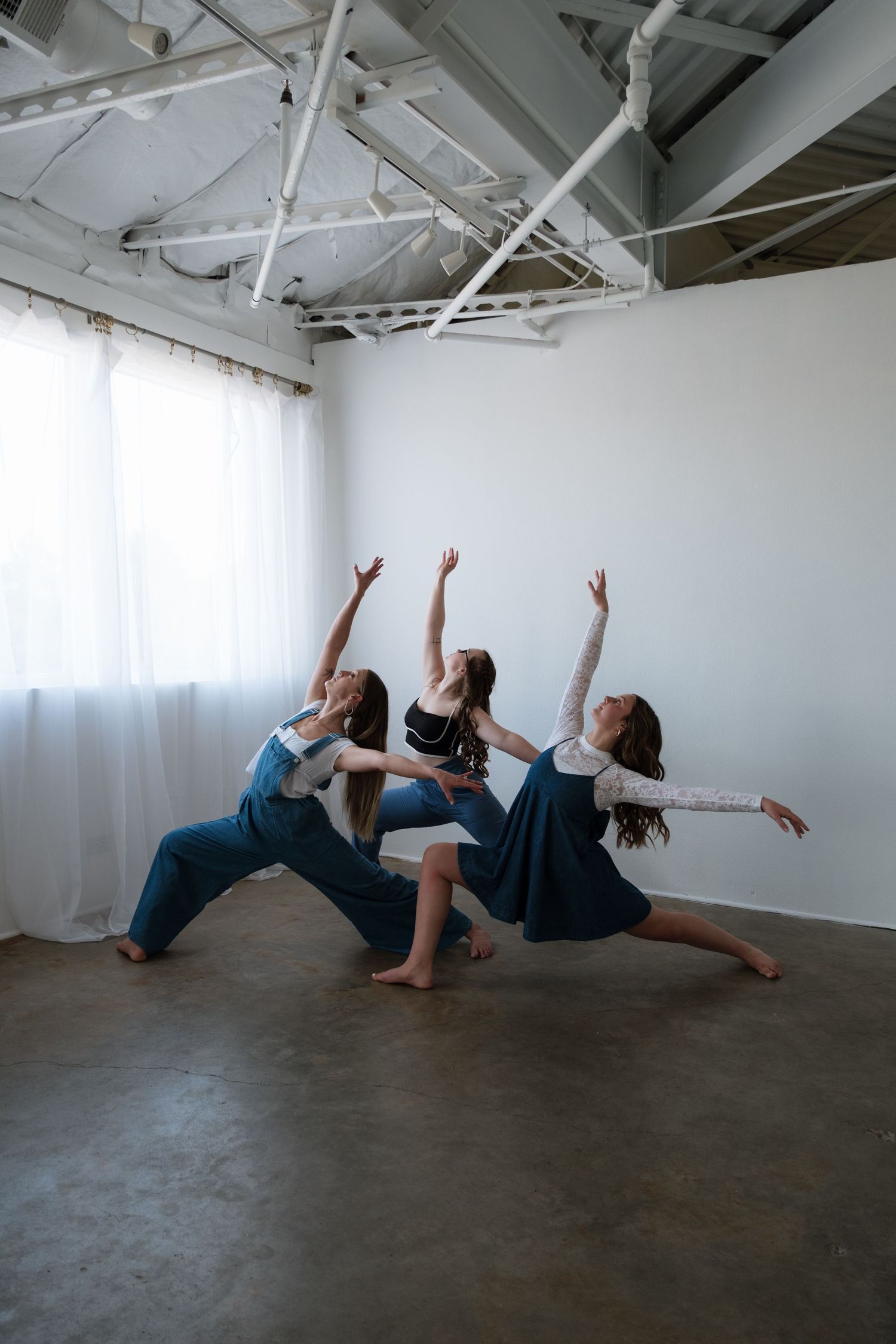 Three female contemporary dancers perform a synchronized pose with arms raised in a bright studio space.