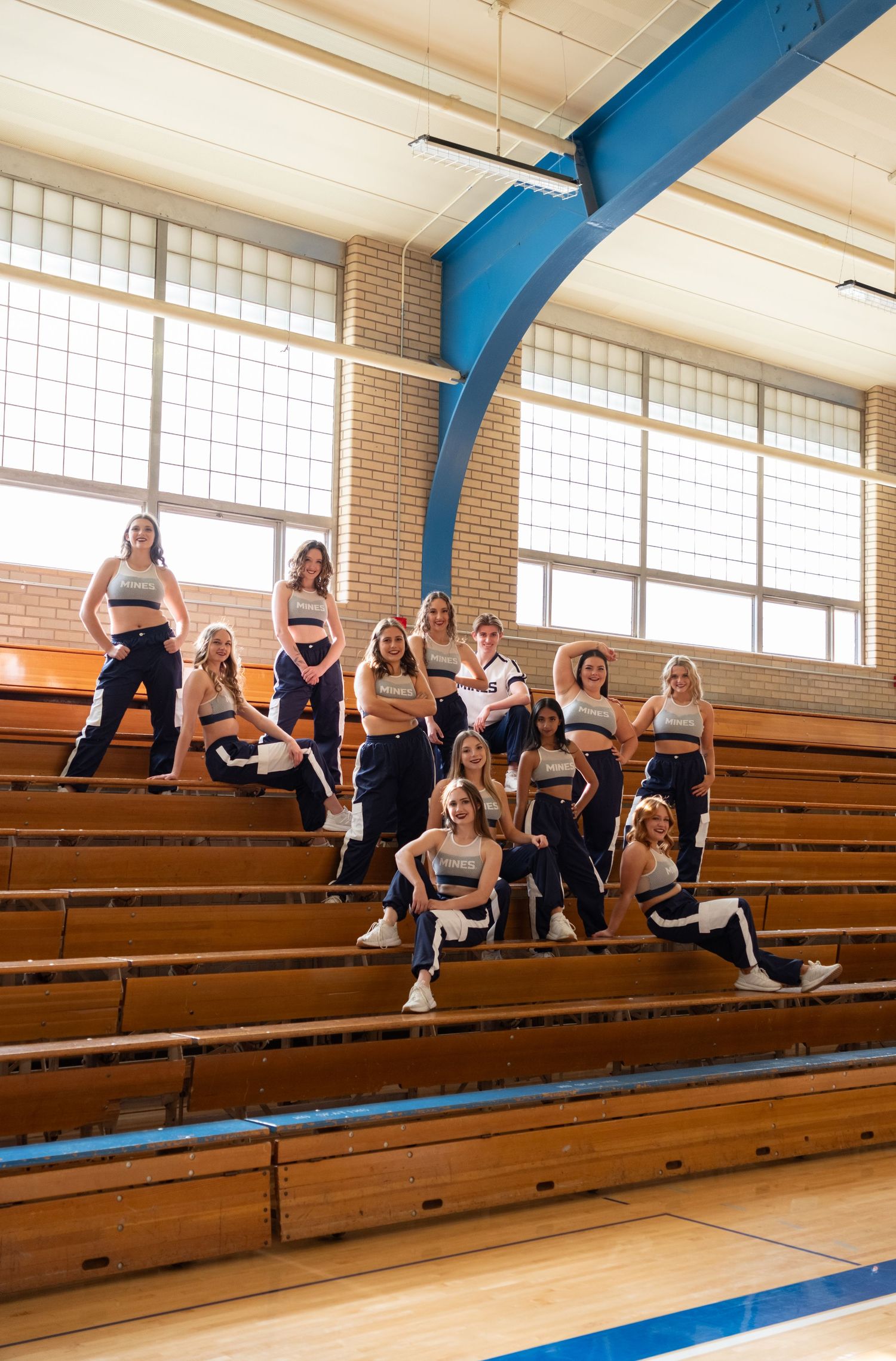 Cheerleaders in navy and white uniforms pose on wooden bleachers inside a school gymnasium.