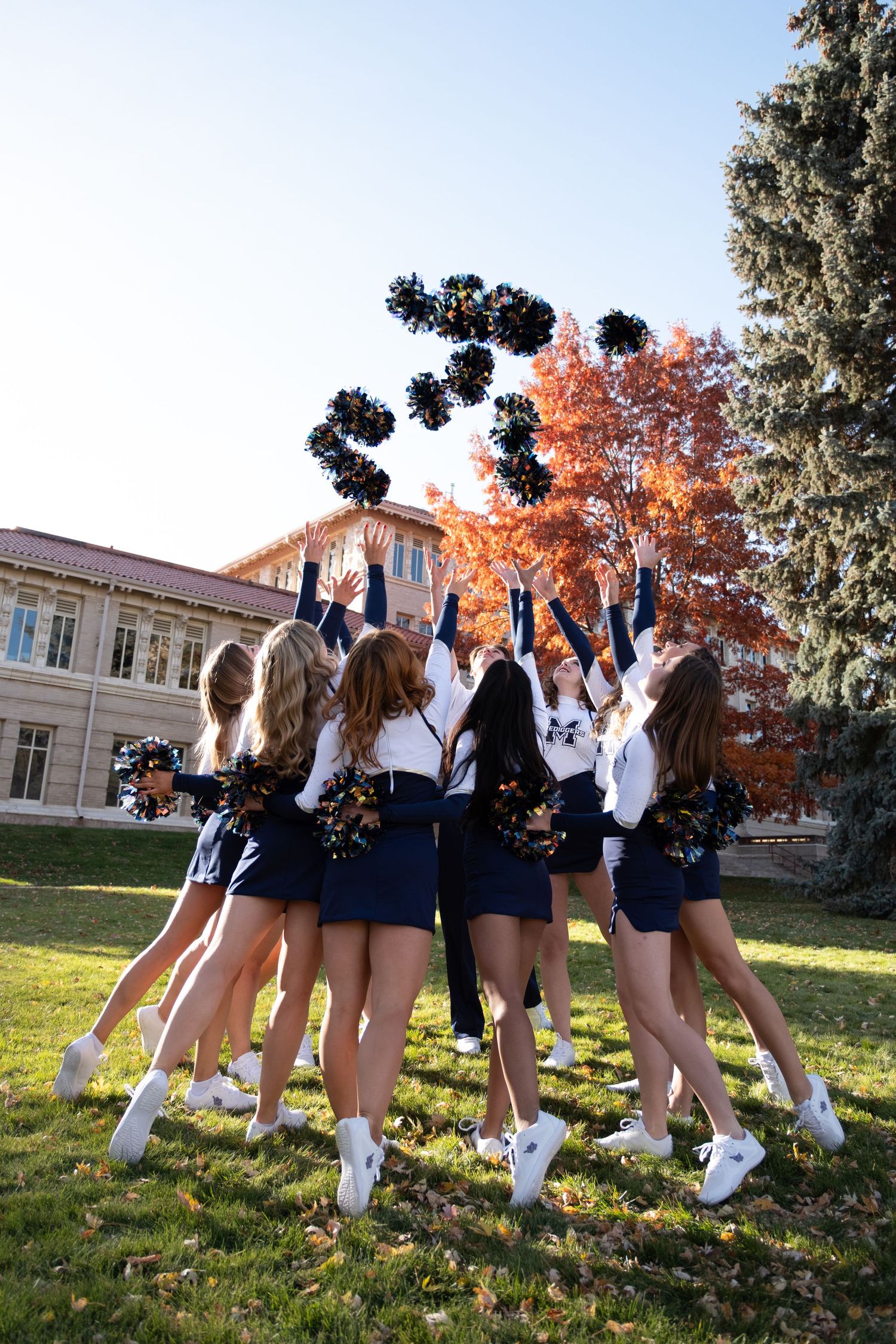 Cheerleaders in navy and white uniforms toss black pom-poms in the air outdoors near a building with autumn trees.