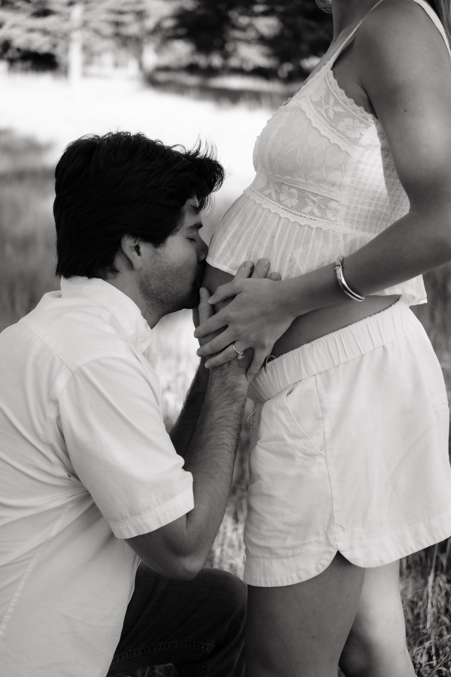 Black and white photo of a man kneeling in a grassy field and kissing the baby bump of his pregnant partner in white lace top and shorts.