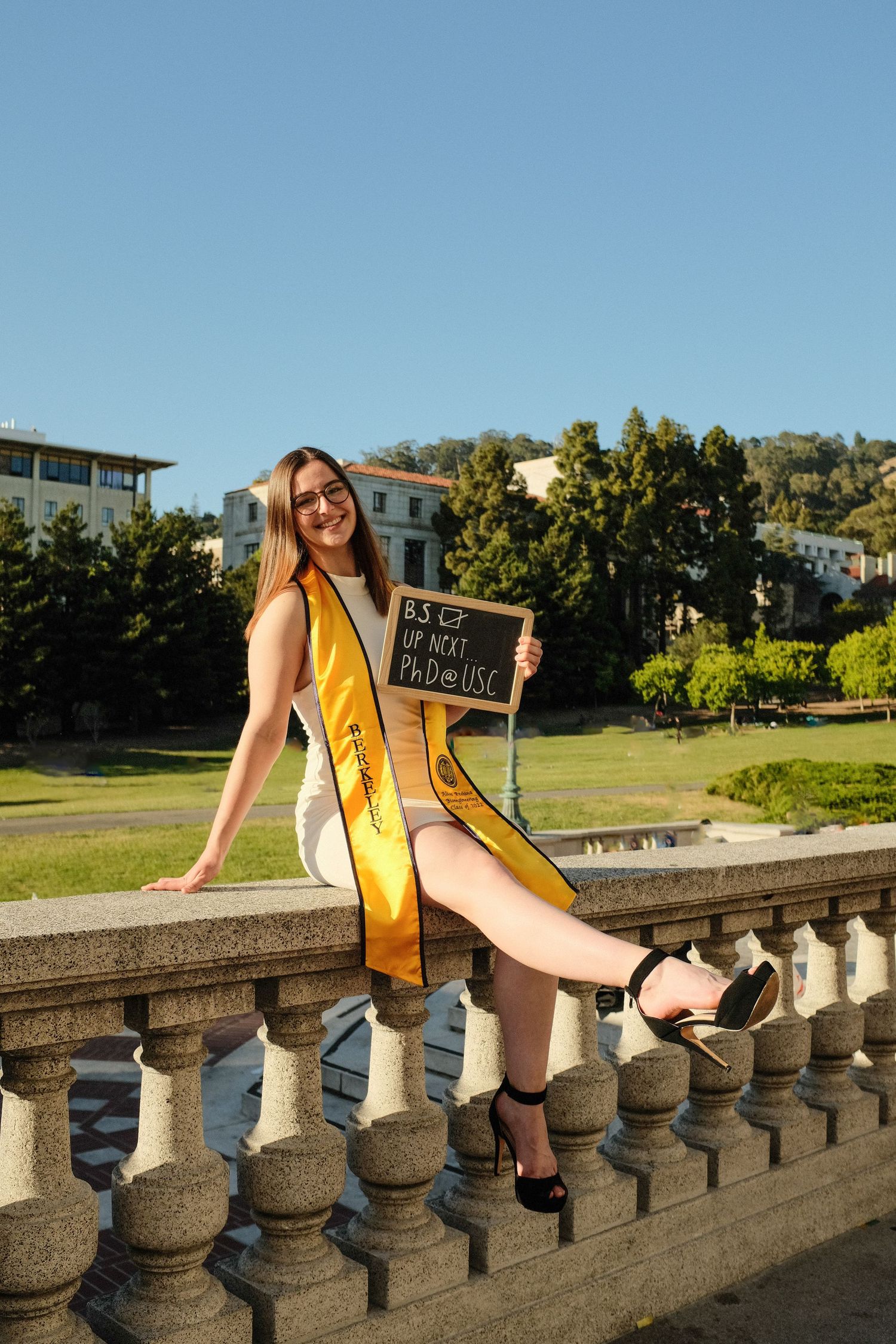 Graduate in yellow stole sits on stone balustrade during outdoor photo session.