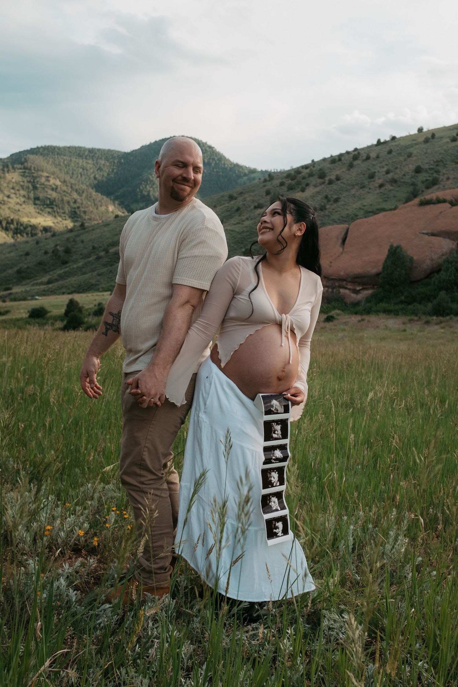 A couple poses for a maternity photo in a grassy field with red rocks and mountains in the background.