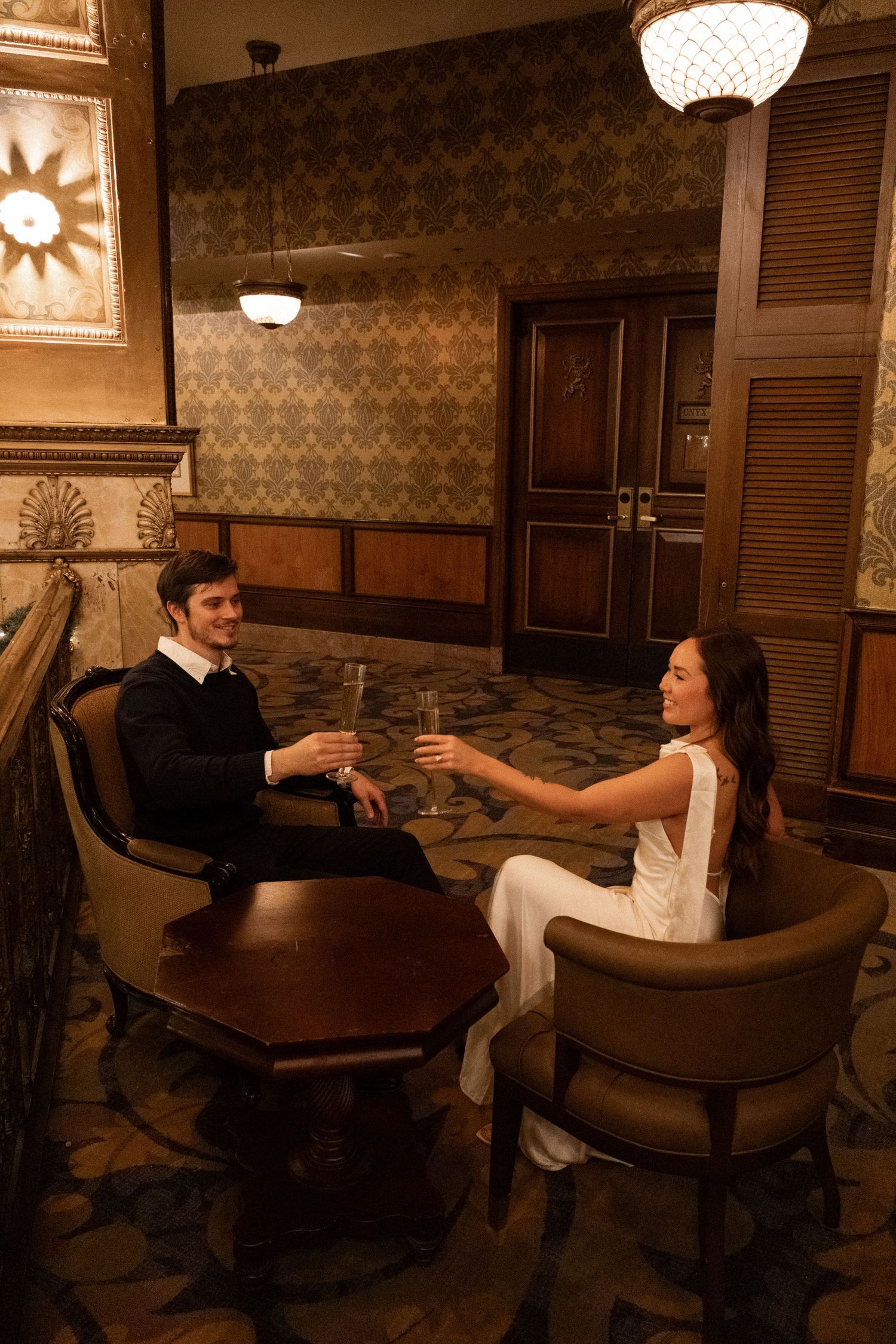 Couple in formal attire toasting champagne glasses at an elegant vintage bar with ornate wallpaper and warm lighting.
