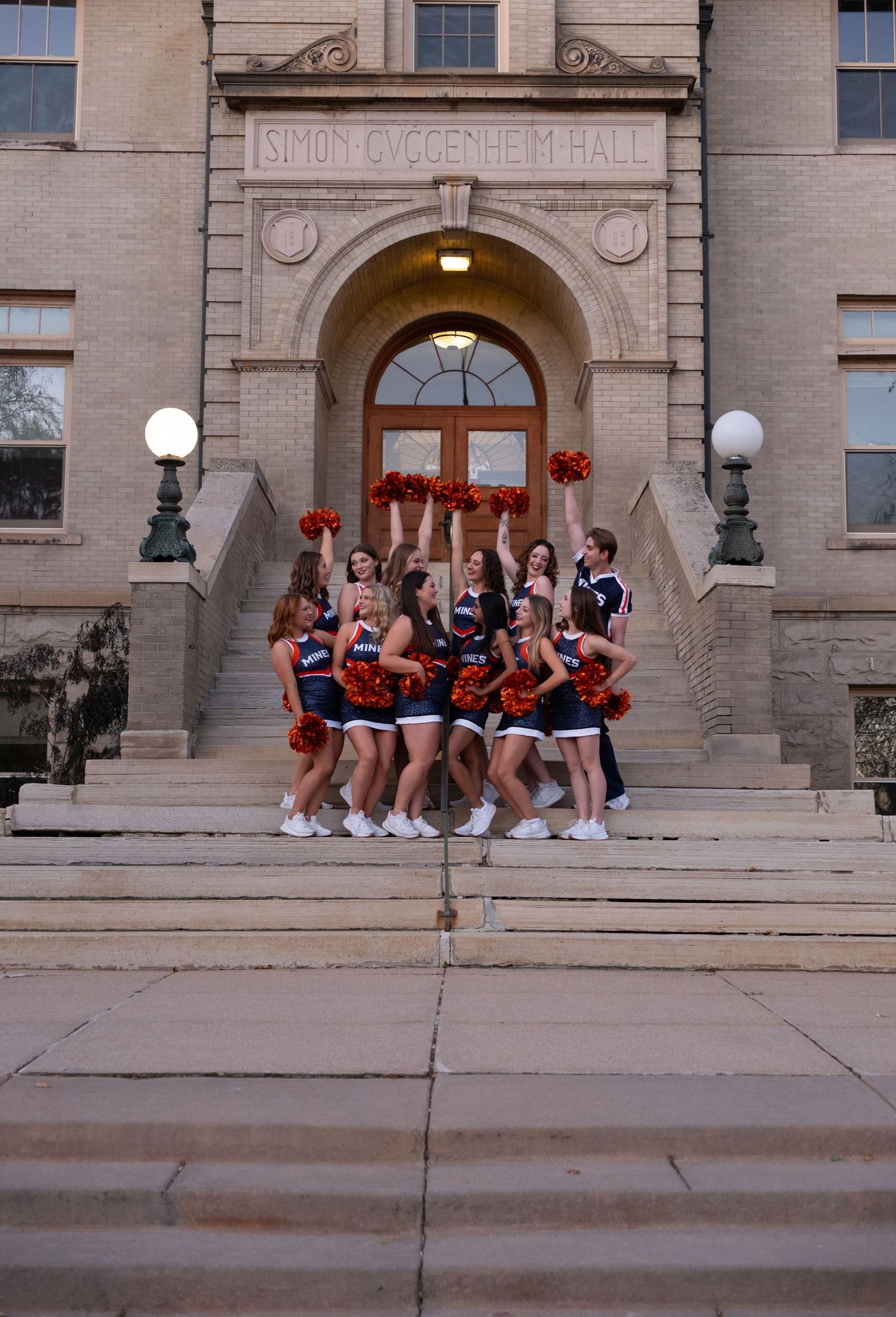 Cheerleaders in blue and orange uniforms with red pom-poms pose on steps of Simon Guggenheim Hall.