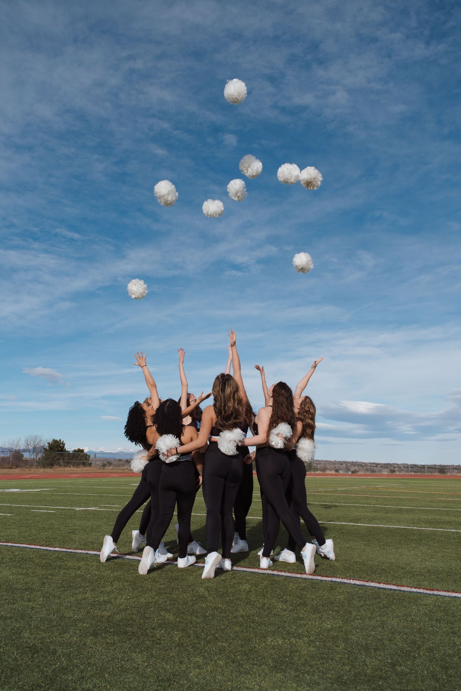 Cheerleaders in black uniforms toss white pom-poms into the sky on a green turf field under a blue cloudy sky.
