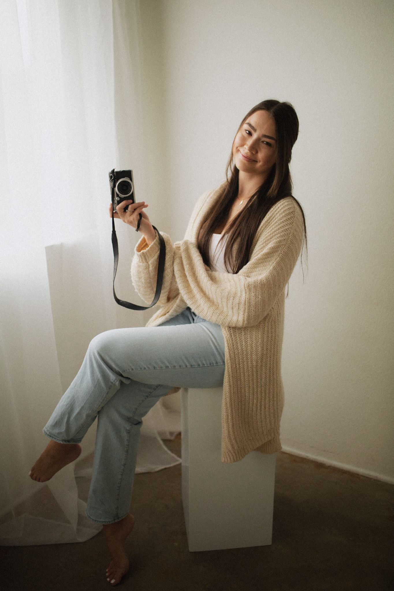 A person in a cream knit sweater and light jeans poses in a minimalist corner setting with a vintage camera.