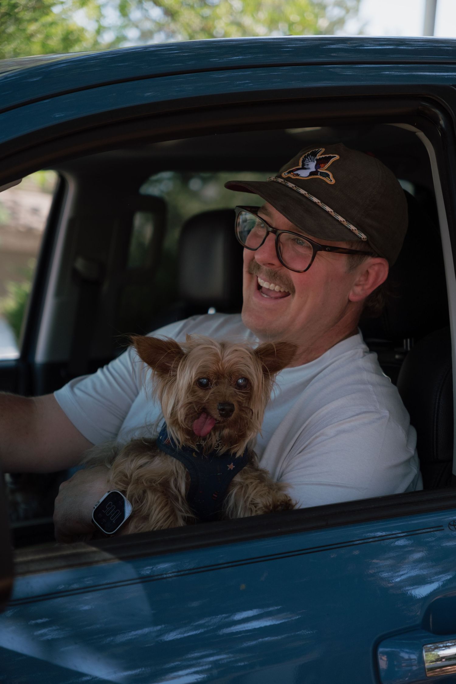 Smiling man in glasses and a baseball cap sits in a driver's seat holding a happy Yorkshire Terrier with its tongue out.