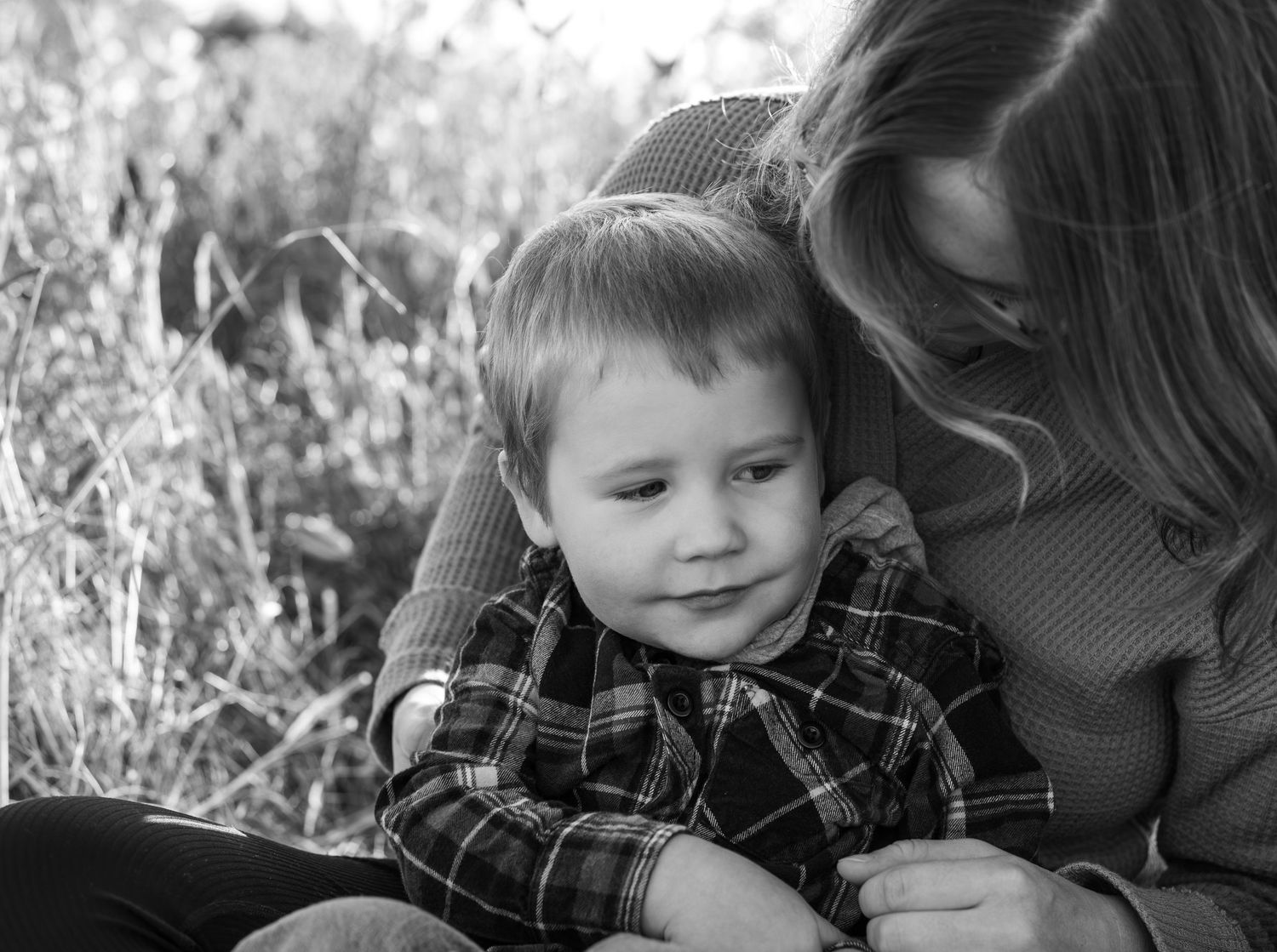 Black and white photo of a young boy in plaid shirt snuggling with his mother outdoors in a field.