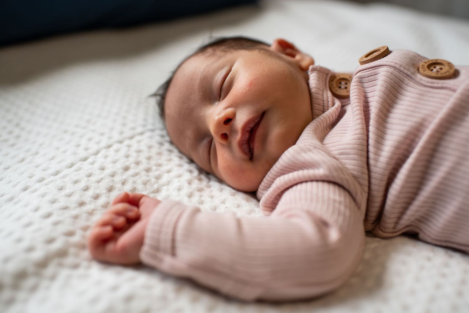 Sleeping newborn in soft pink sweater rests peacefully on white textured bedding.
