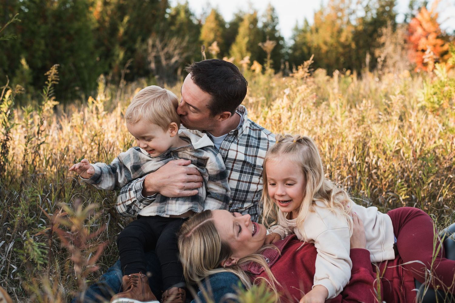 Family of four enjoying a warm autumn day in a golden meadow, with parents and two young children laughing together.