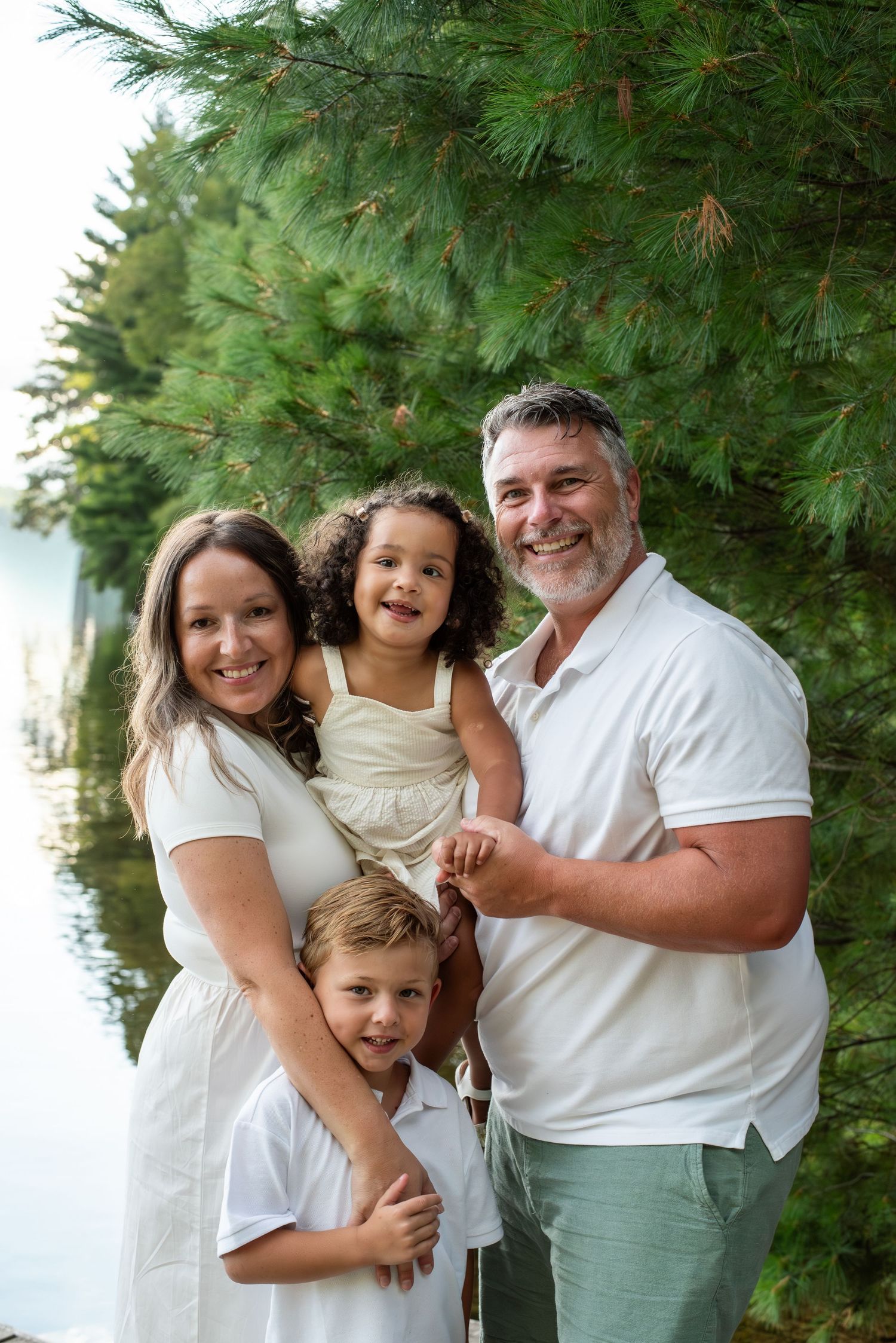 Happy family of four posing outdoors near a lake surrounded by lush green pine trees on a summer day.