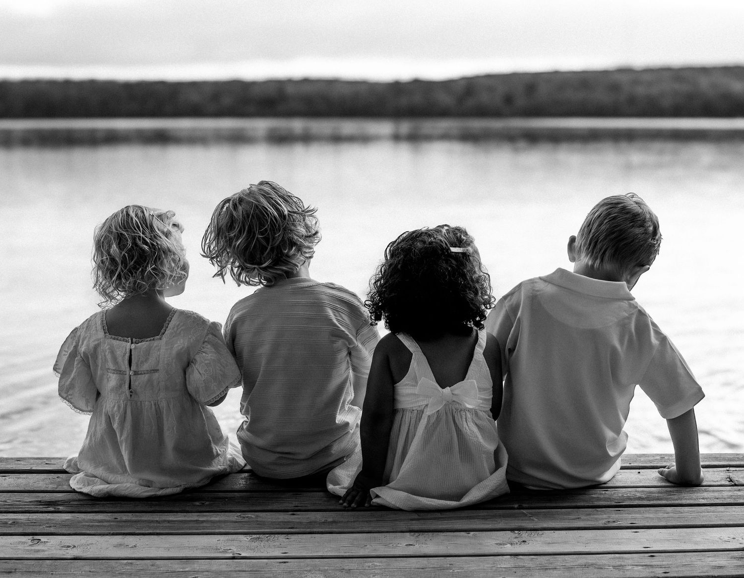 Four children sit together on a wooden dock overlooking a calm lake in a black and white photo.
