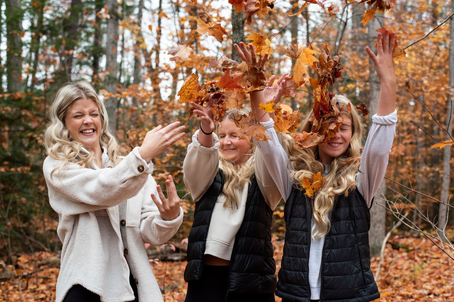 Three women joyfully tossing autumn leaves in a forest, surrounded by golden fall foliage.