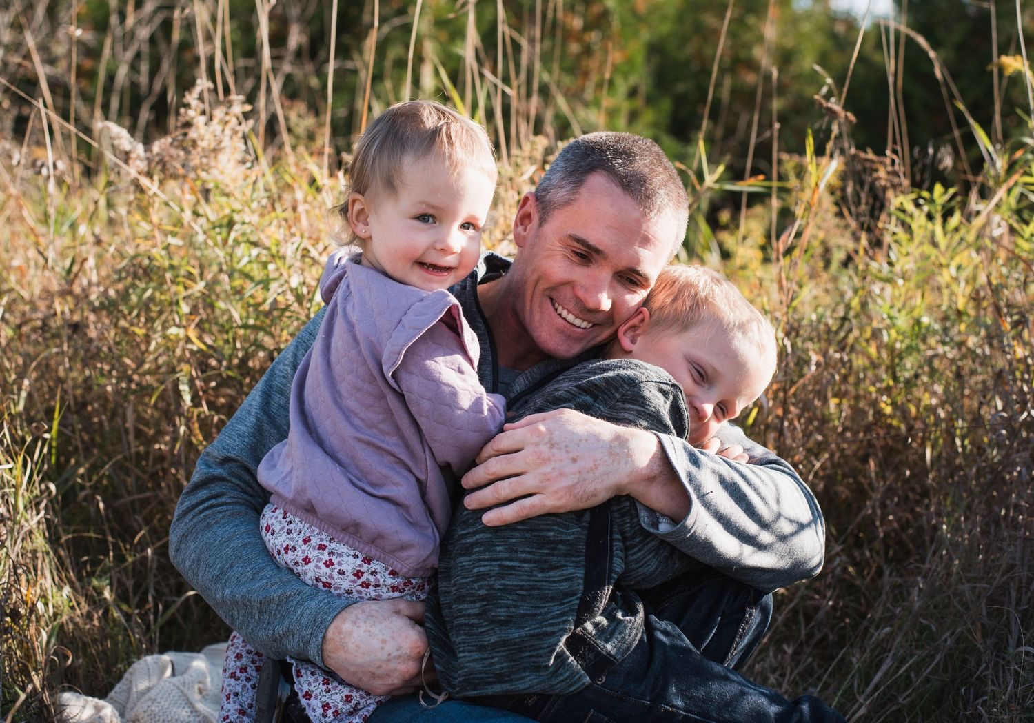 A smiling man sits outdoors hugging two young children in a grassy field on a sunny day.