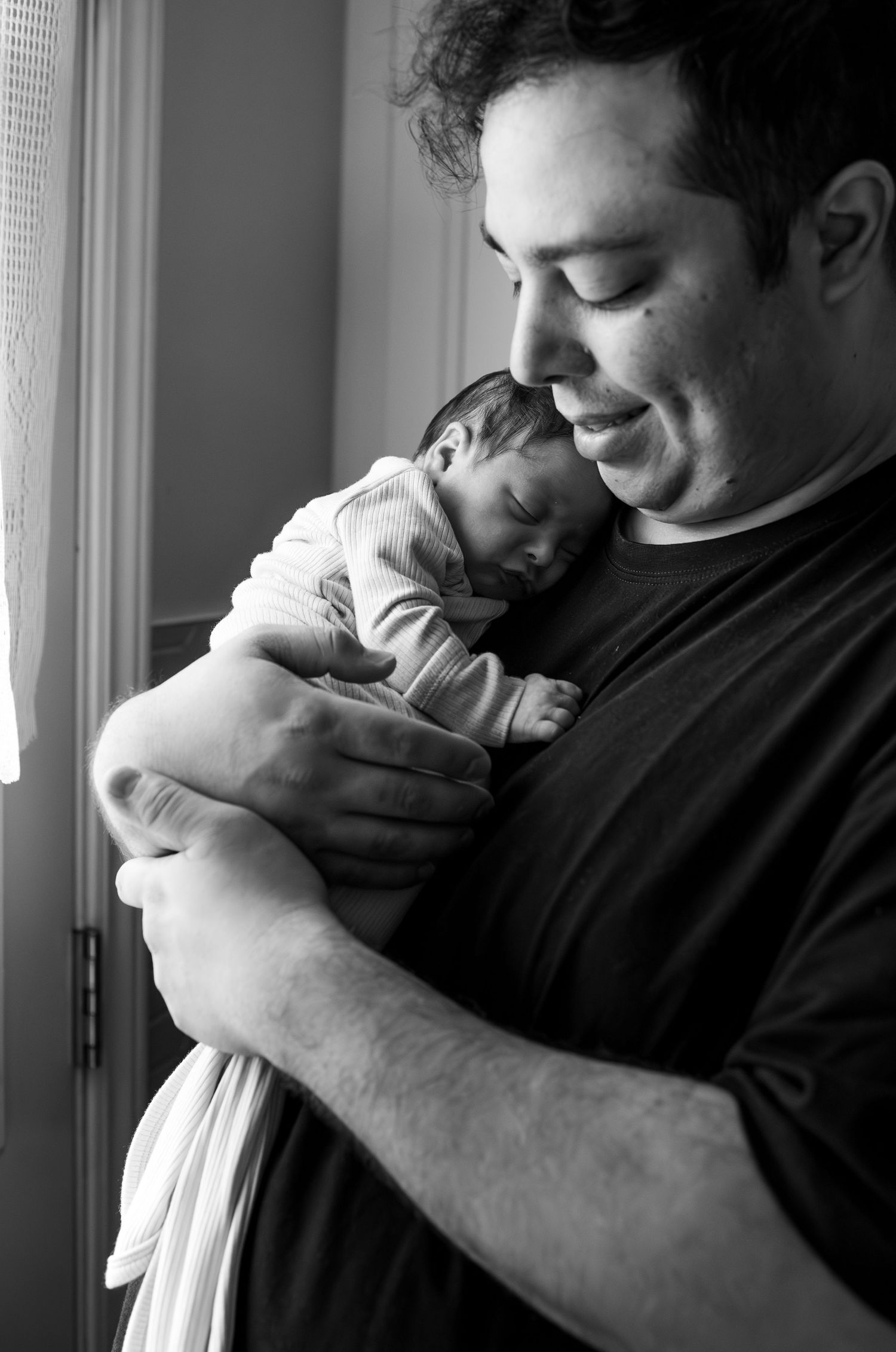 Black and white portrait of someone cradling a sleeping newborn baby while standing by a window.