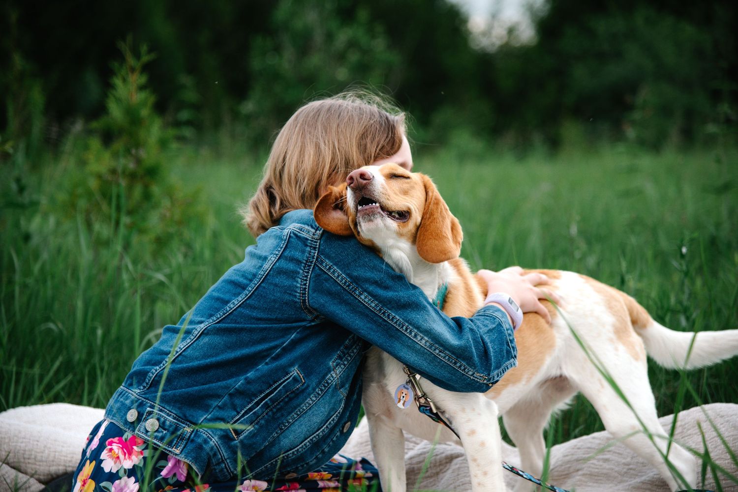 A young girl in a denim jacket hugs a beagle dog outdoors in a lush green field.
