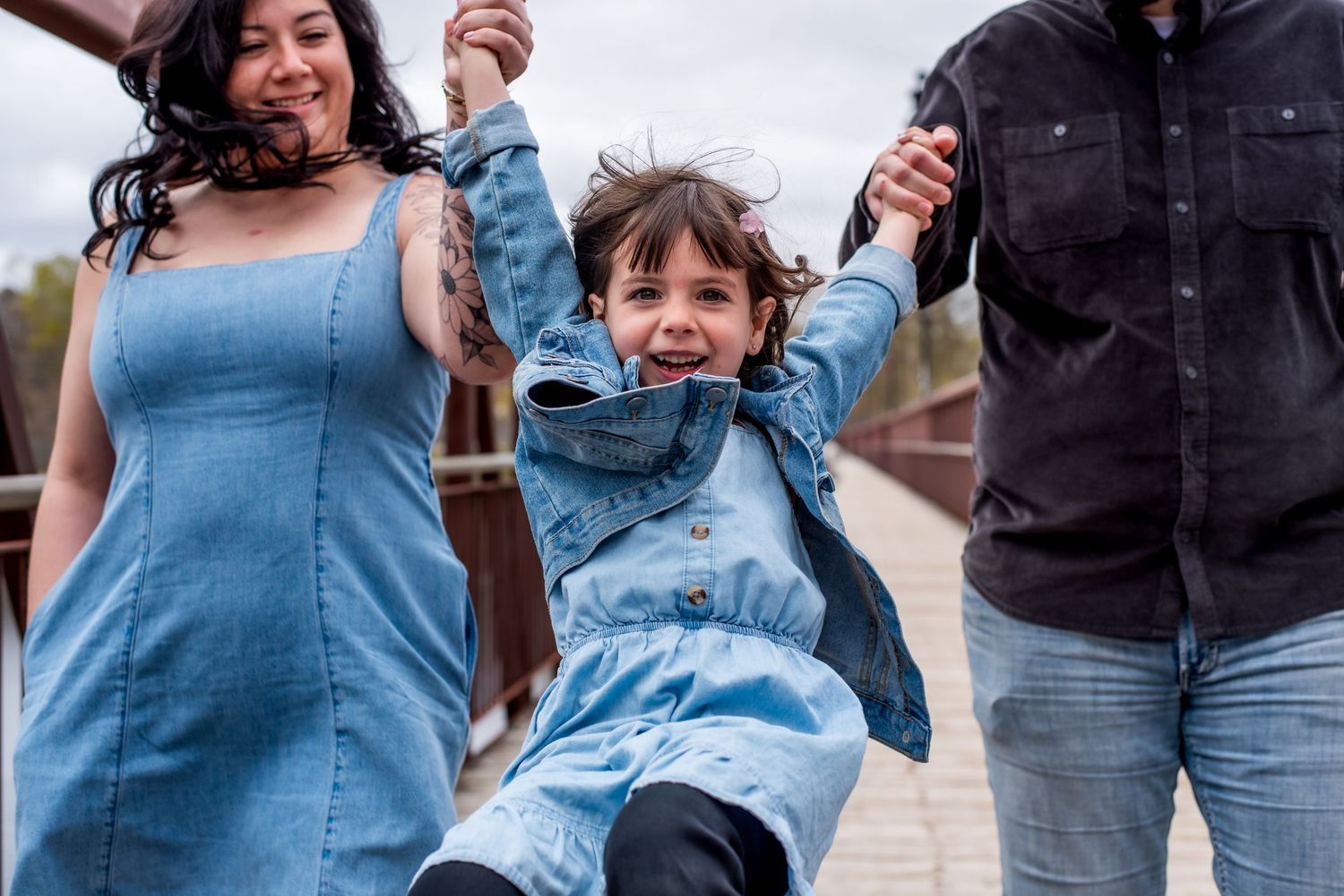 A joyful family swings their young daughter between them while walking on a bridge, all wearing denim outfits.