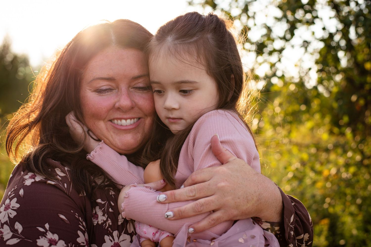 A smiling woman hugs a young girl outdoors in golden sunlight, both with dark hair, wearing pink and floral outfits.