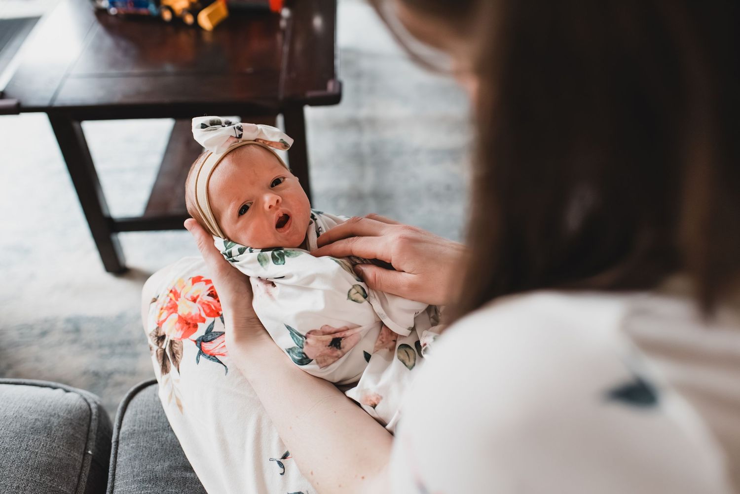 A person feeding a baby wrapped in a floral blanket while sitting on a gray couch.