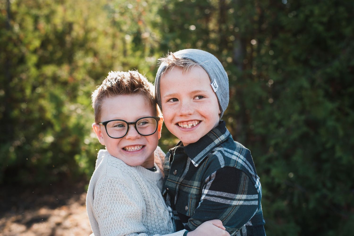 Two children smile together outdoors wearing glasses and a beanie in natural lighting.