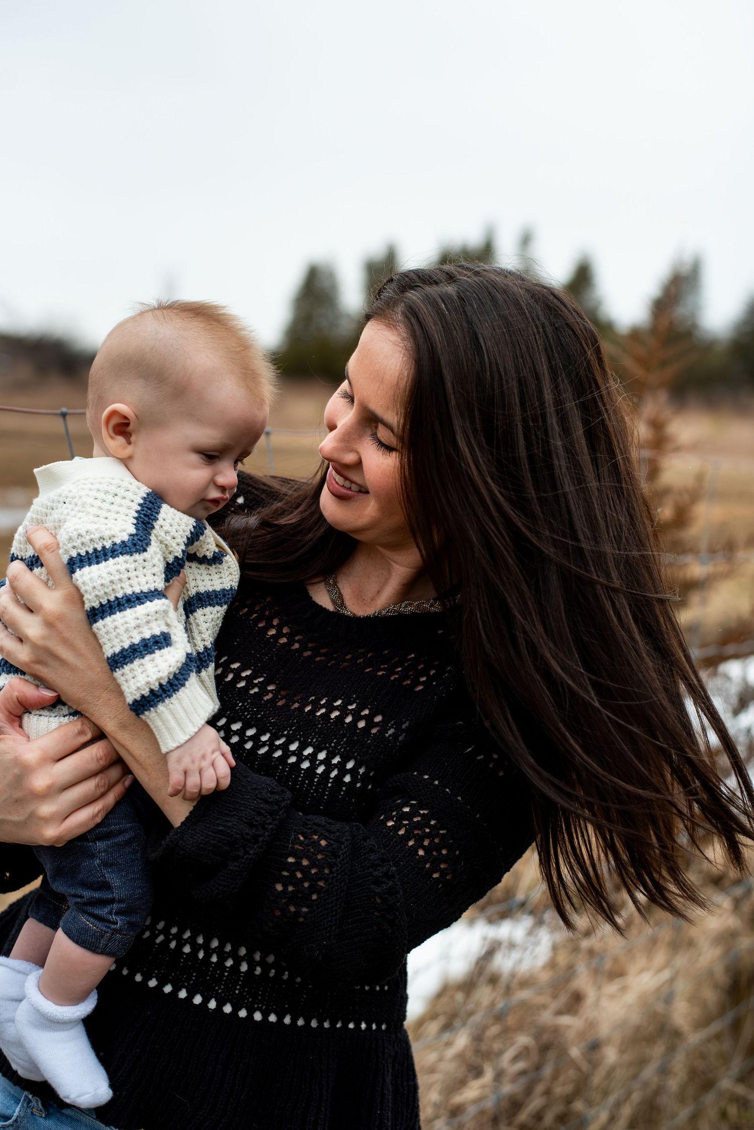 A mother and baby share a tender moment outdoors, wearing matching striped sweaters against a natural backdrop.